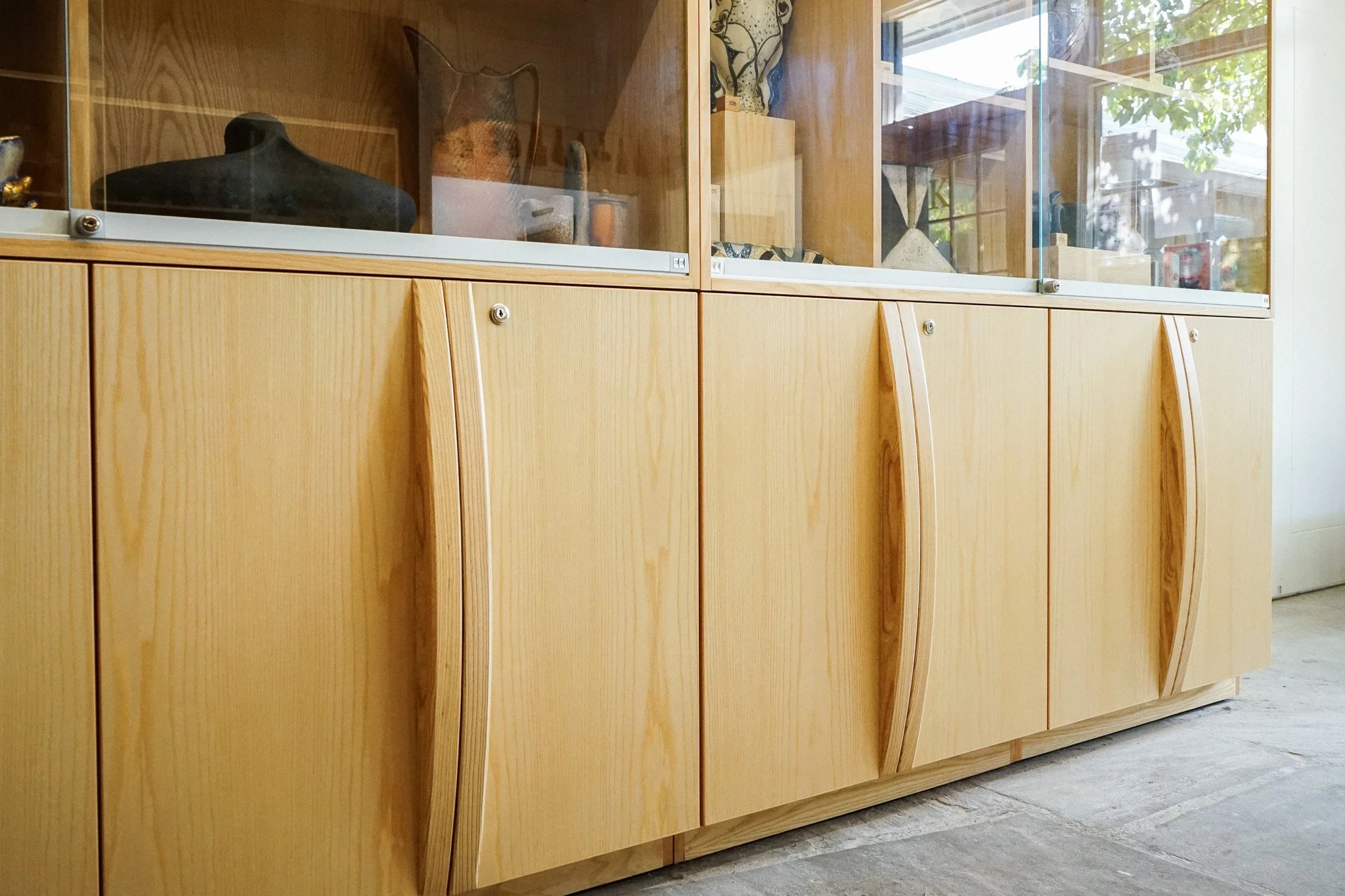 A wooden display cabinet with glass doors containing various vases and ceramic objects. The cabinet has a natural finish with curved design elements on the lower doors, and it is situated near a window showing outside greenery.
