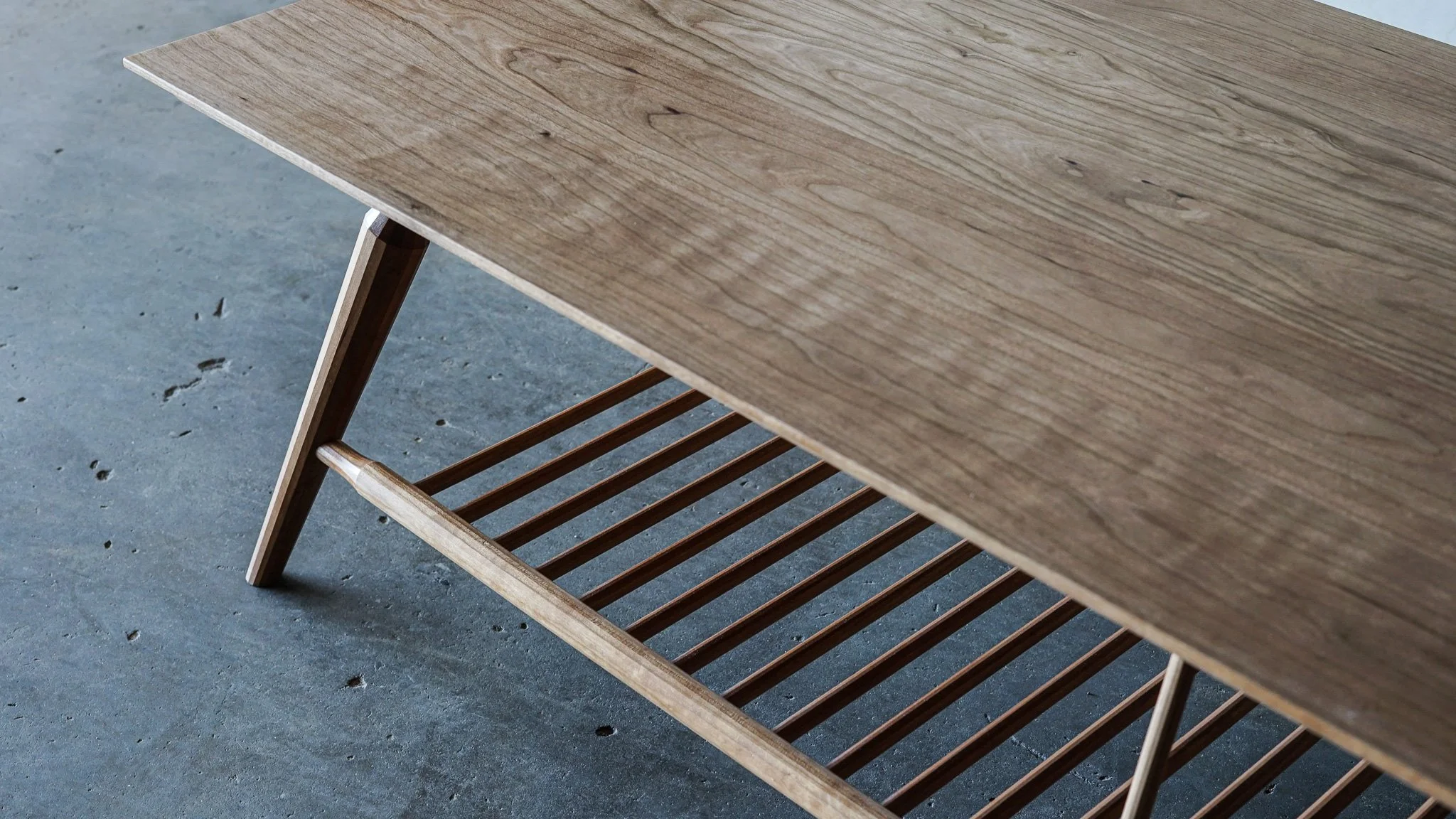 A detail of a cherry wooden coffee table with angled legs and a slatted shelf underneath, placed on a concrete floor.