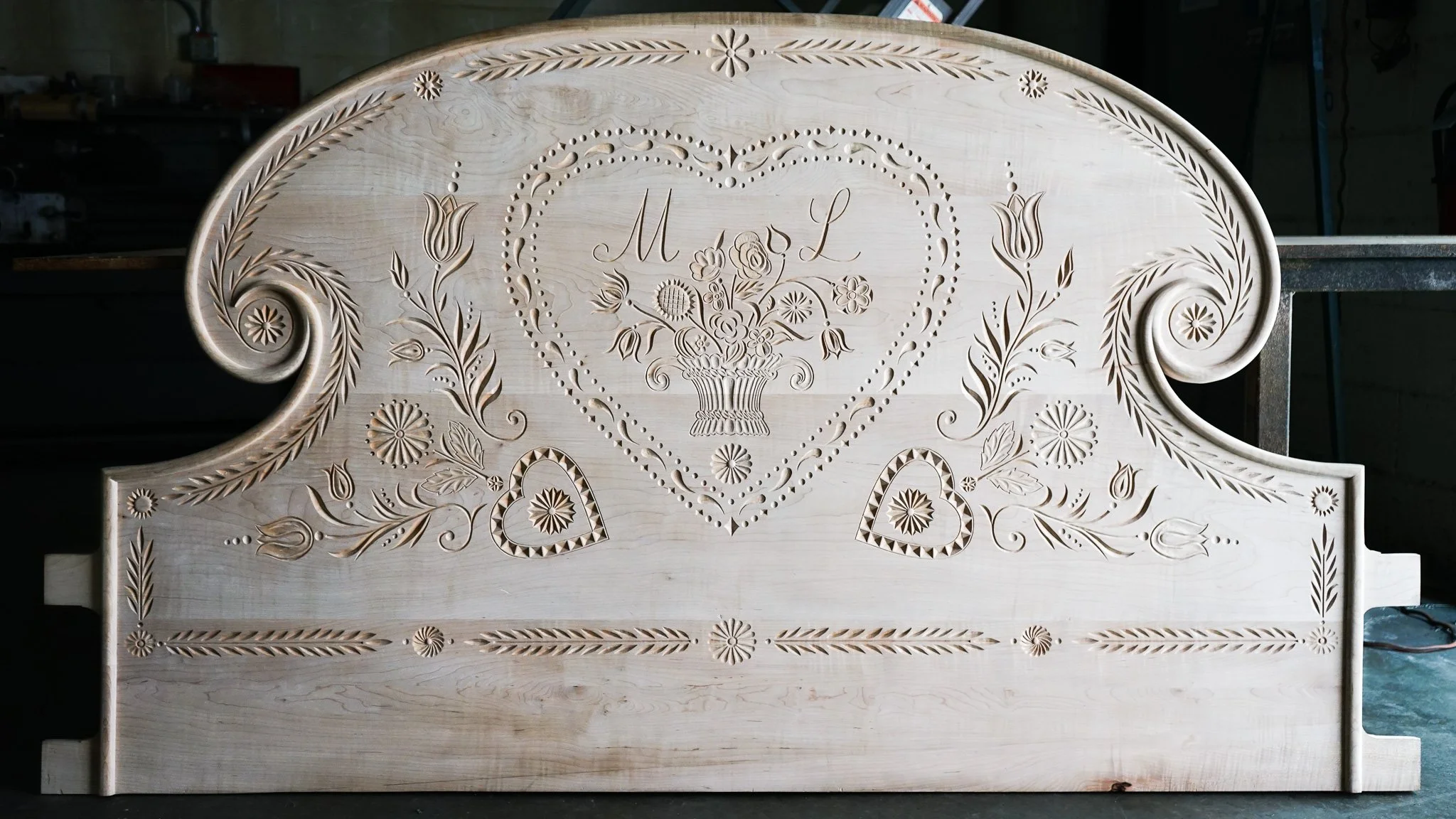 A wooden headboard with carved floral and heart patterns, featuring initials 'M L' in the center, in a workshop or garage setting.
