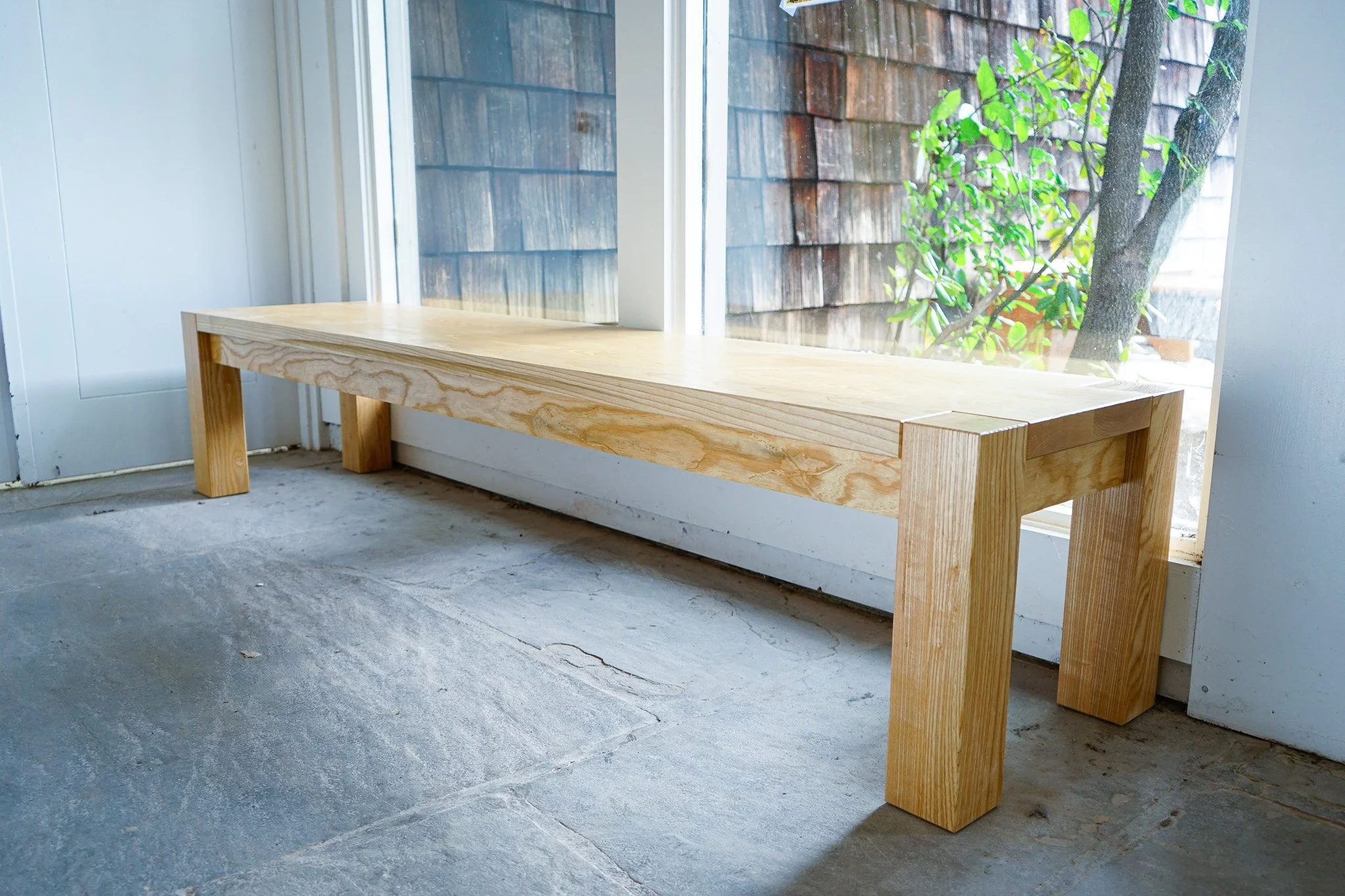 A wooden bench with a simple, sturdy design positioned in front of a window. The window reveals a view of a outdoor wooden fence and a green-leafed tree.