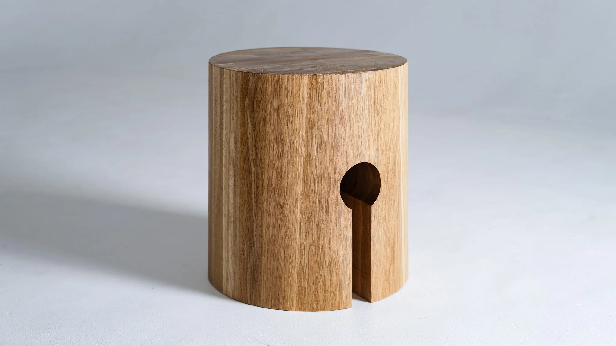 A cylindrical wooden side table with a hole on the side, made of white oak wood, against a plain light gray background.