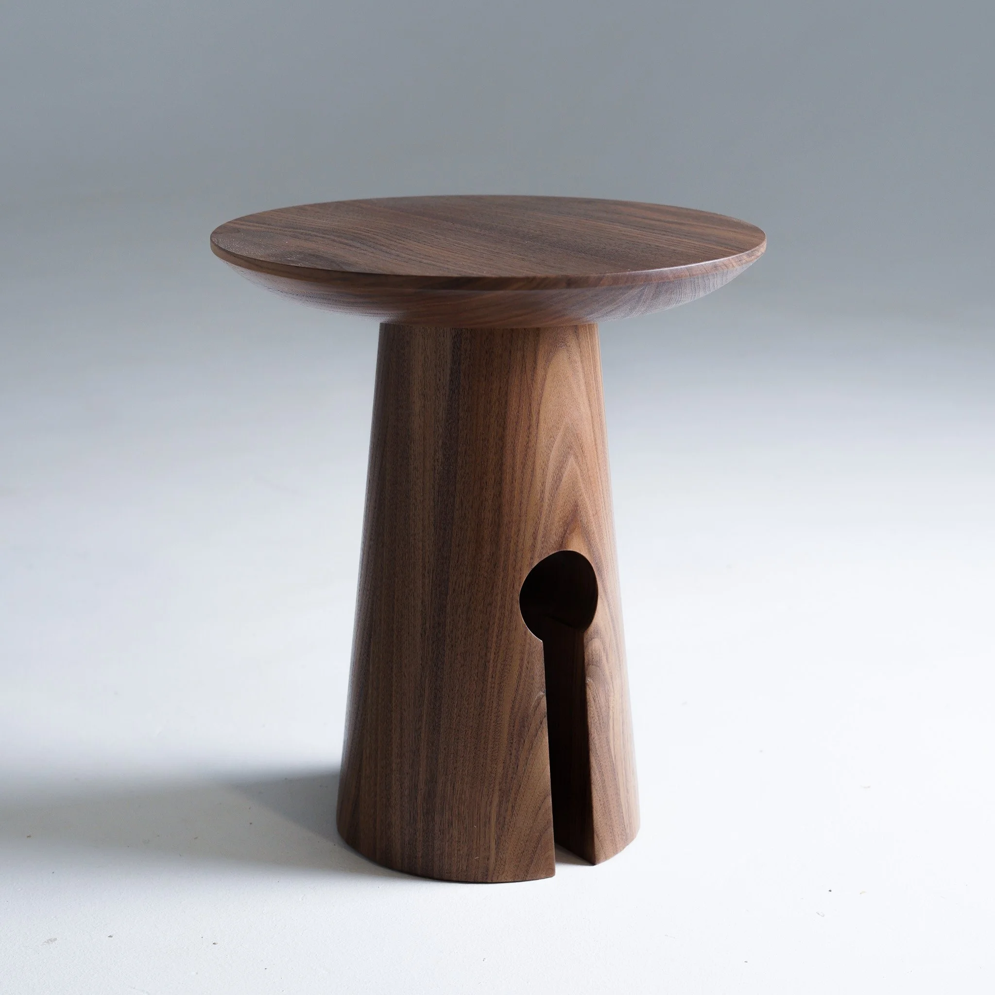 A round wooden side table with a thicker top and a pedestal base, made of walnut wood with visible grain patterns, placed on a light-colored surface against a plain gray background.