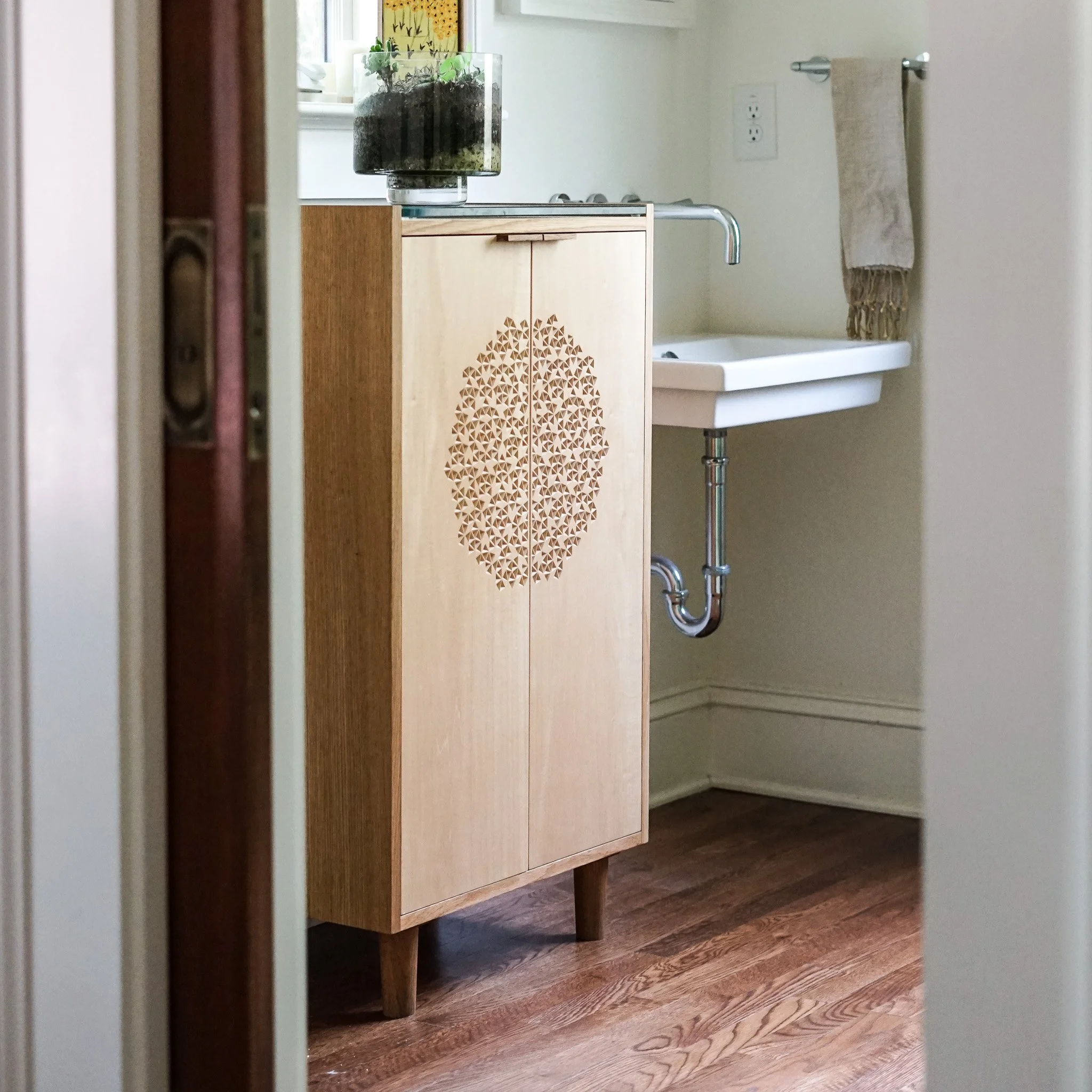 A wooden vanity cabinet with decorative geometric pattern on the doors, standing on a hardwood floor in a bathroom. A small white sink is partially visible behind the cabinet, and a beige towel hangs on a horizontal bar on the wall to the right.