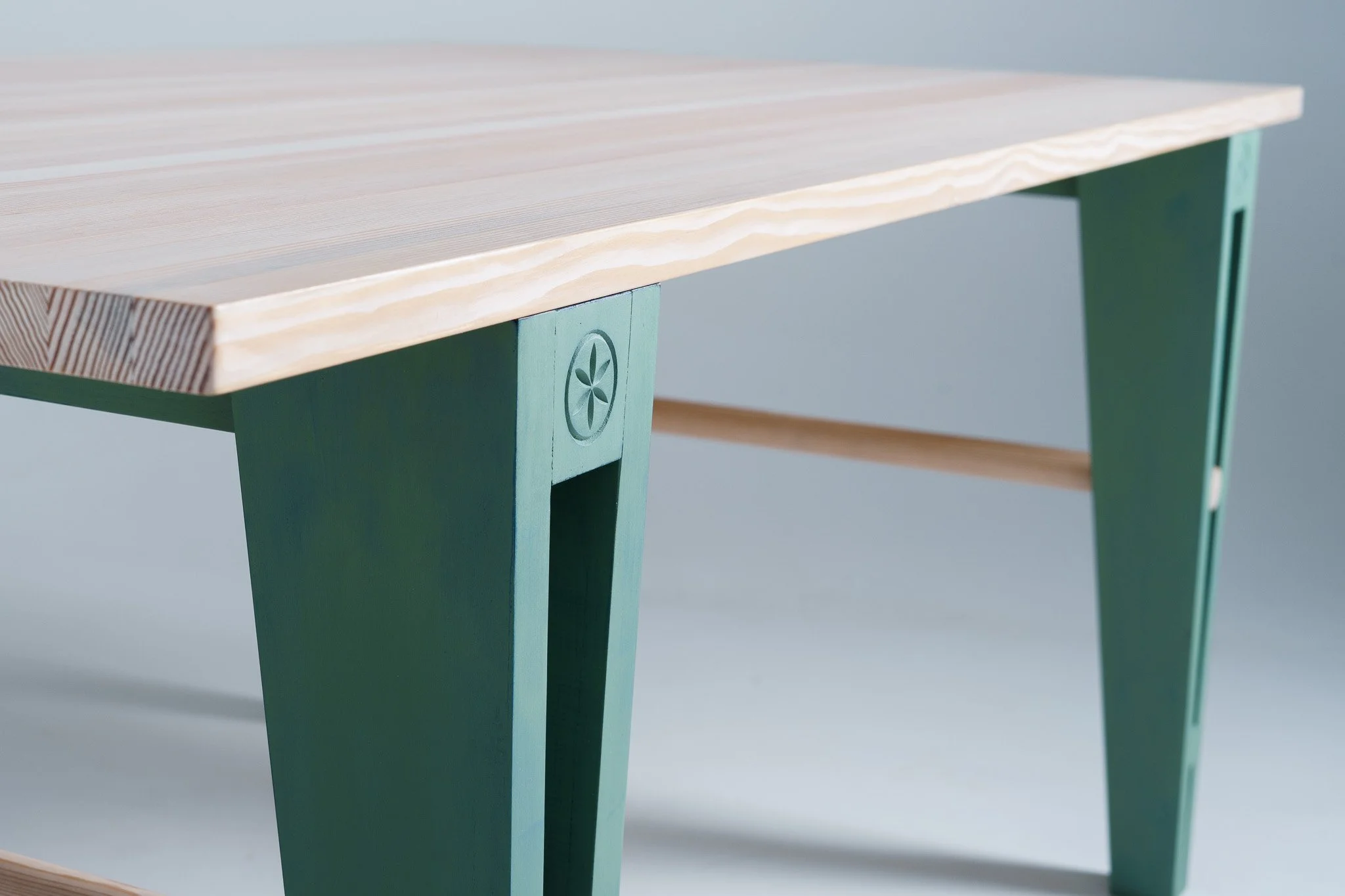 Close-up of a wooden table with a light-colored pine top and green painted legs, one of which has a carved flower emblem.