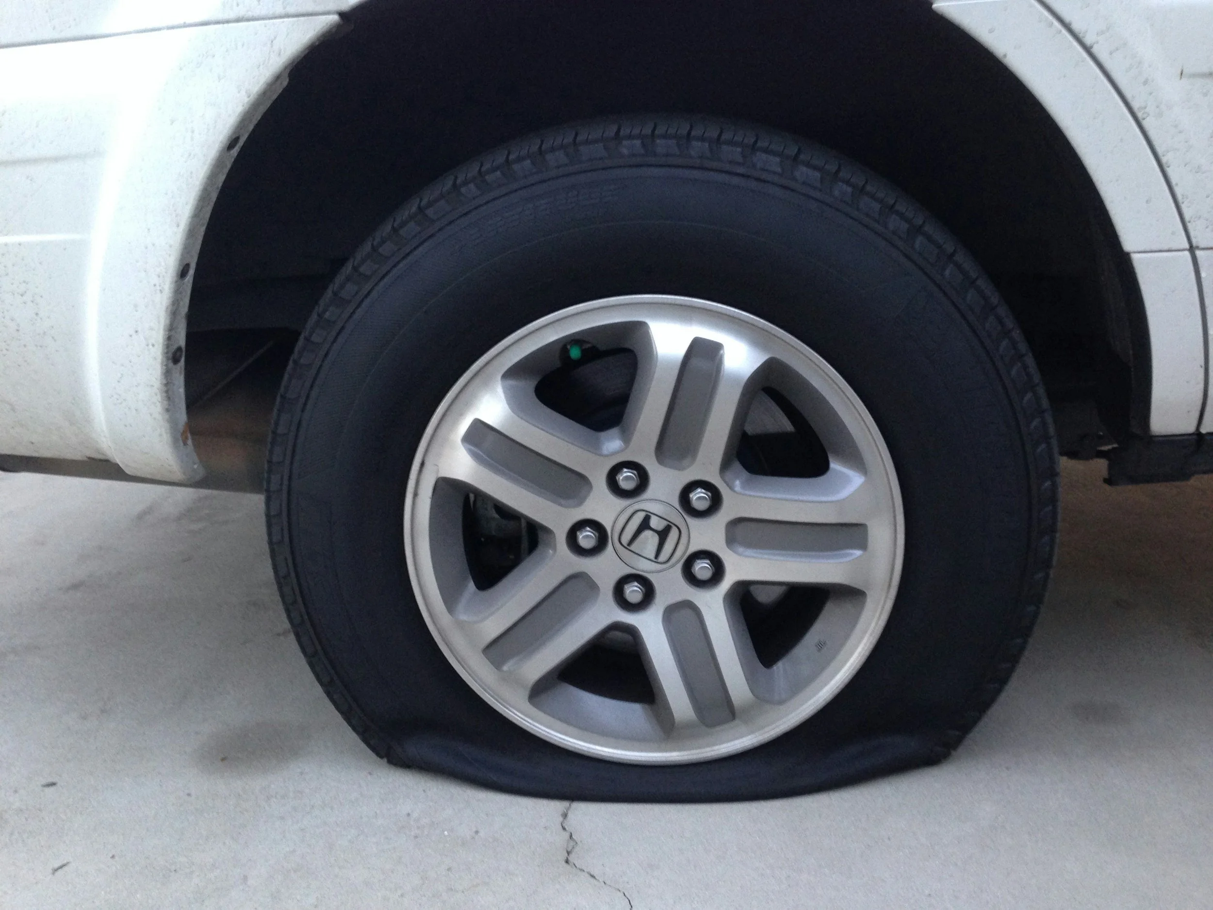 Close-up of a car wheel with a black tire and silver alloy rim, showing the Honda logo at the center, mounted on a white vehicle parked on a concrete surface.