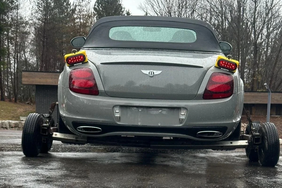 A silver Bentley convertible car with removed rear wheels, showing the car mounted on a wheel-less chassis, parked outdoors on a wet surface with trees and a wooden railing in the background.