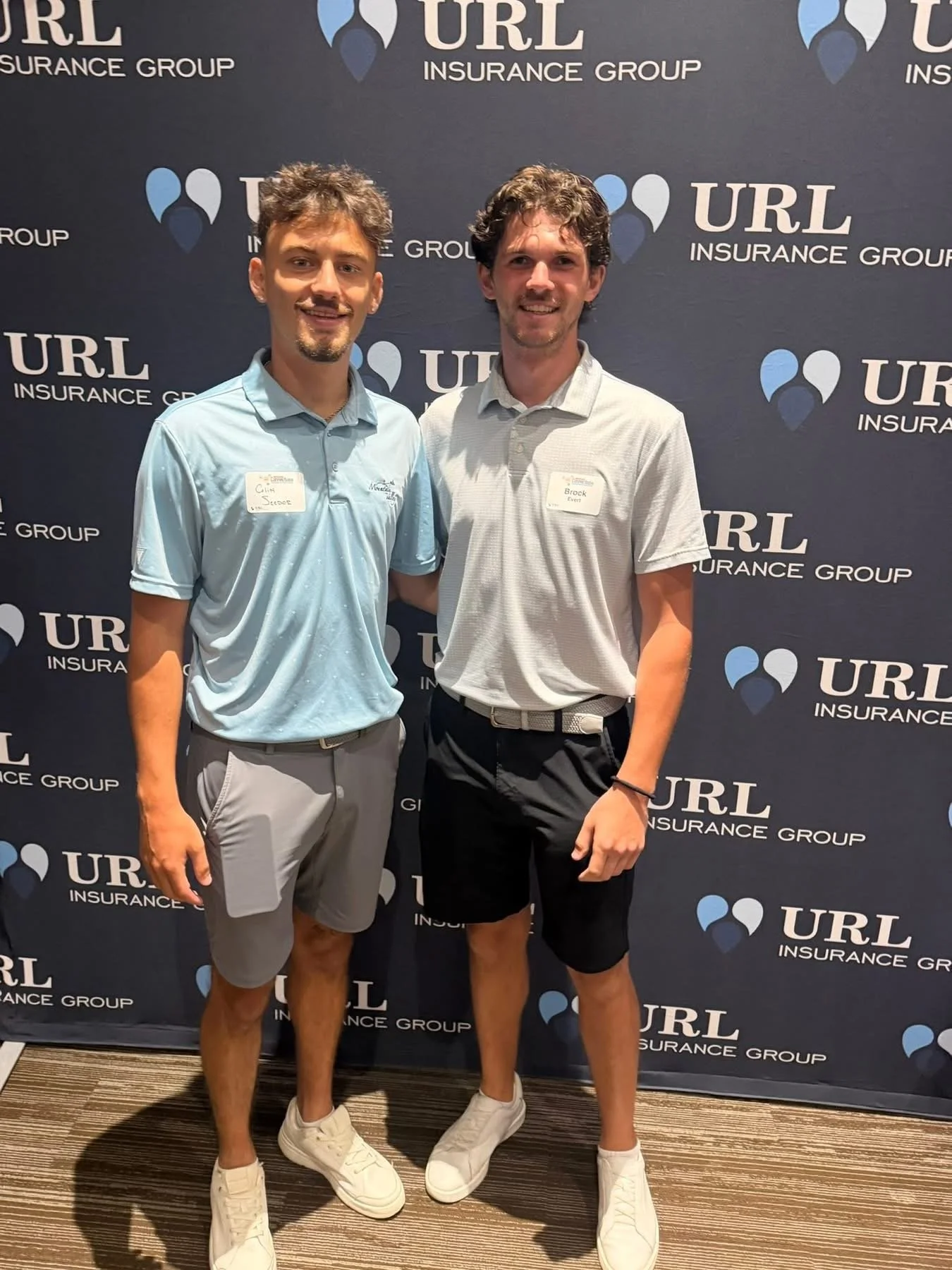 Two young men standing in front of a dark blue backdrop that displays the logo and name of URL Insurance Group. They are smiling, wearing casual sports attire, and have name tags on their shirts.
