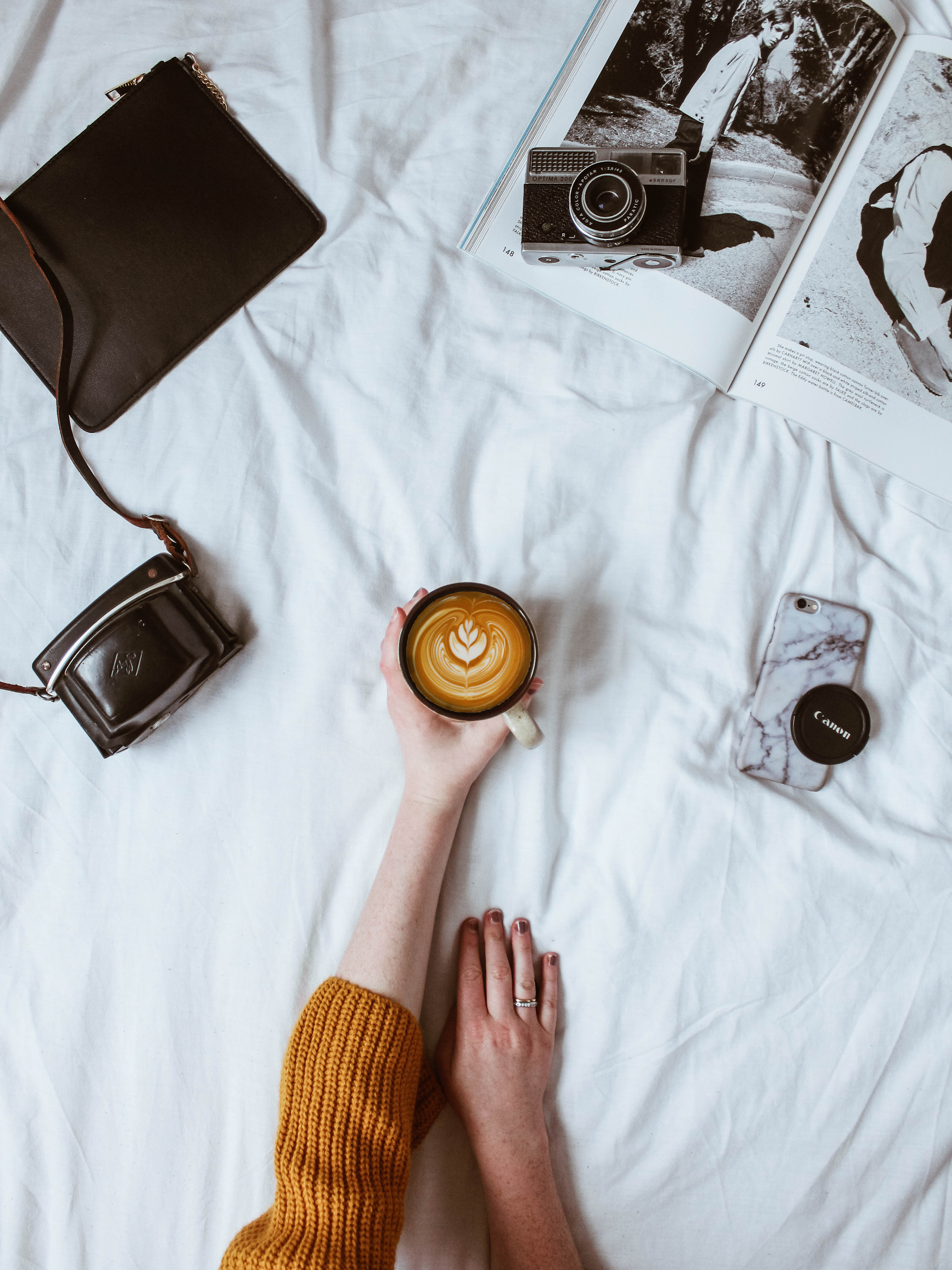 Overhead view of a person with a yellow sweater holding a coffee cup with latte art, surrounded by a camera, magazines, a cell phone, a handbag, and a camera case on a white bed sheet.