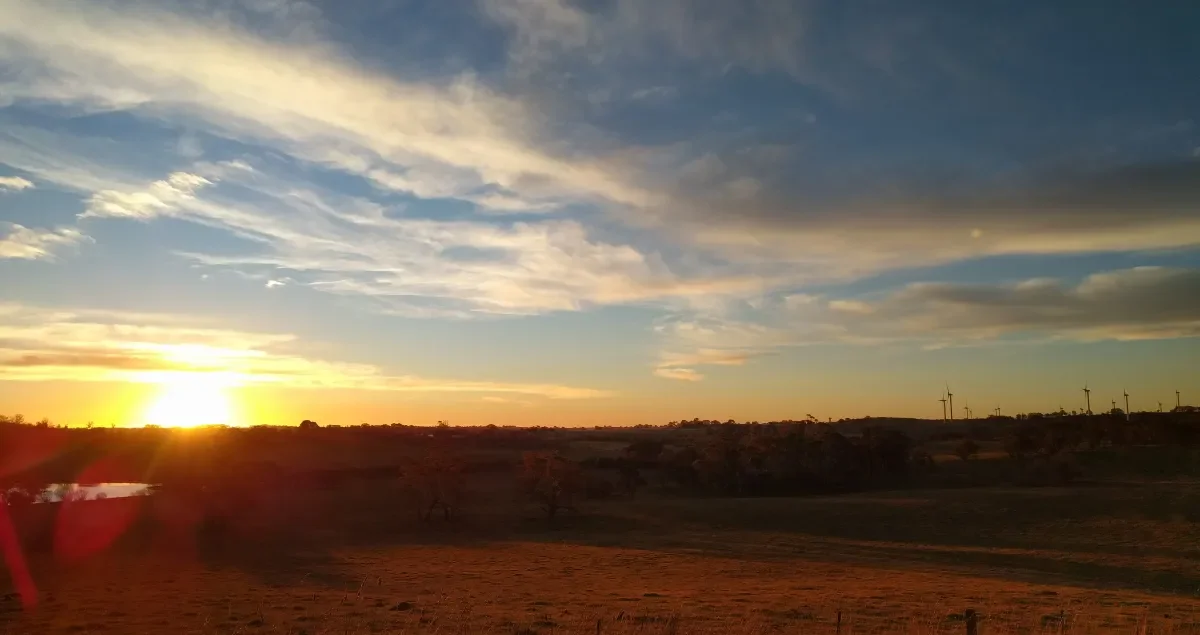Sunset over a rural landscape with wind turbines in the distance and a partly cloudy sky.
