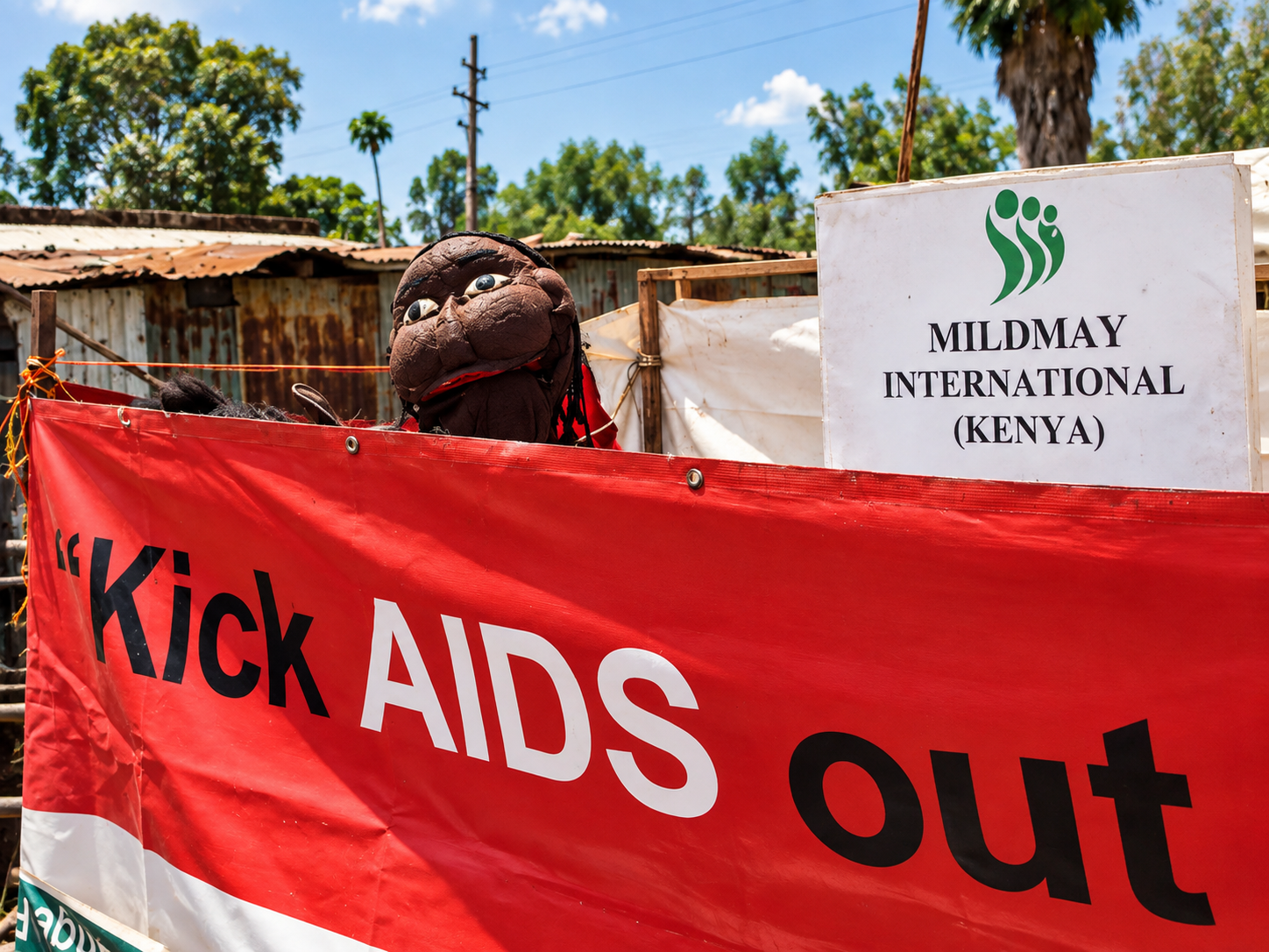 A puppet with a smiling face and large eyes is seen peeking over a red banner that says 'Kick AIDS out' at the Mildmay International Kenya event, with trees, power lines, and a rusty roof in the background.