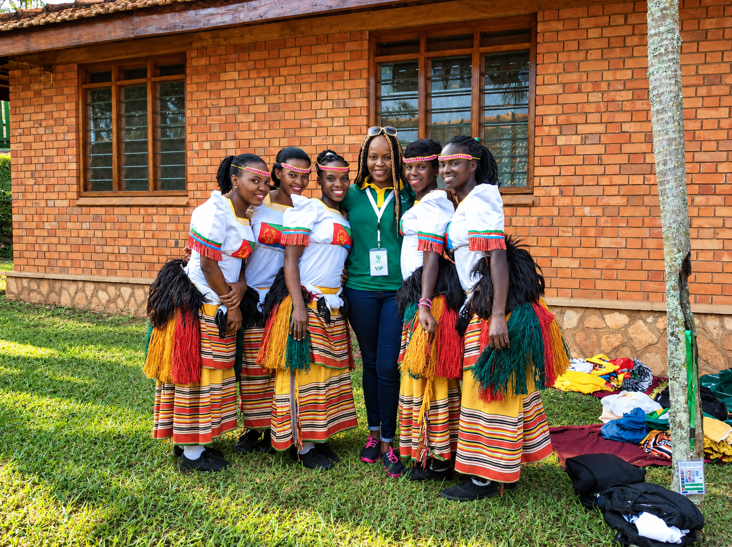A group of six women, five dressed in traditional African attire with colorful skirts, white tops with red and green accents, and black footwear, pose with a woman in casual green and jeans in front of a brick building with wooden windows. They are standing on green grass outdoors.