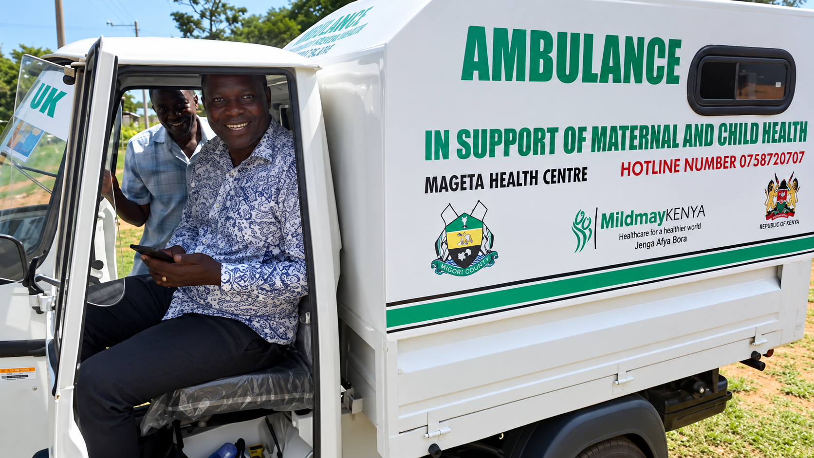 Two men smiling inside a white mobile healthcare vehicle with green text that says 'AMBULANCE IN SUPPORT OF MATERNAL AND CHILD HEALTH' parked on grass outdoors.