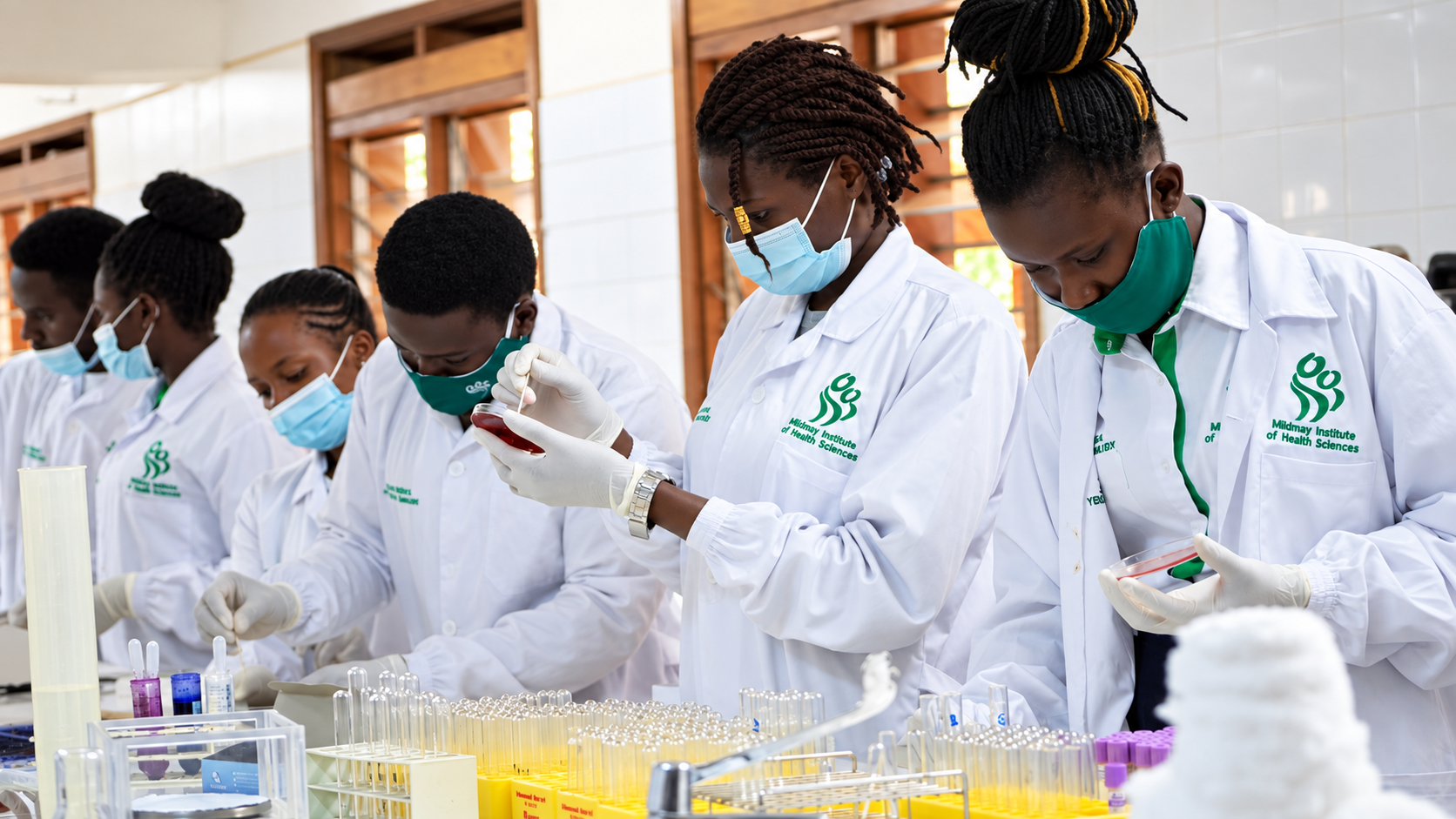 A group of students wearing white lab coats and masks working in a science laboratory, conducting experiments with test tubes and laboratory equipment.
