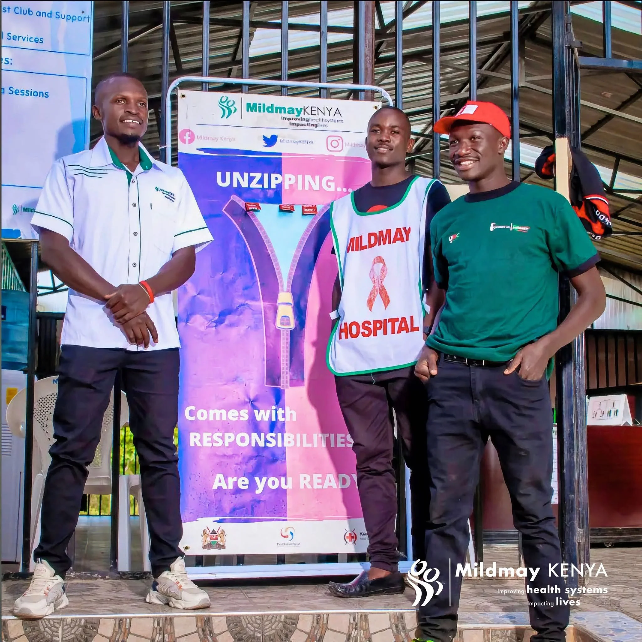 Three men standing outdoors in front of a colorful banner promoting Mildmay Kenya, with the words 'UNZIPPING... Responsibility' and social media icons. The man on the left wears a white shirt with green accents, the man in the middle wears a white apron with red and green, and the man on the right wears a green T-shirt and a red cap, all smiling.