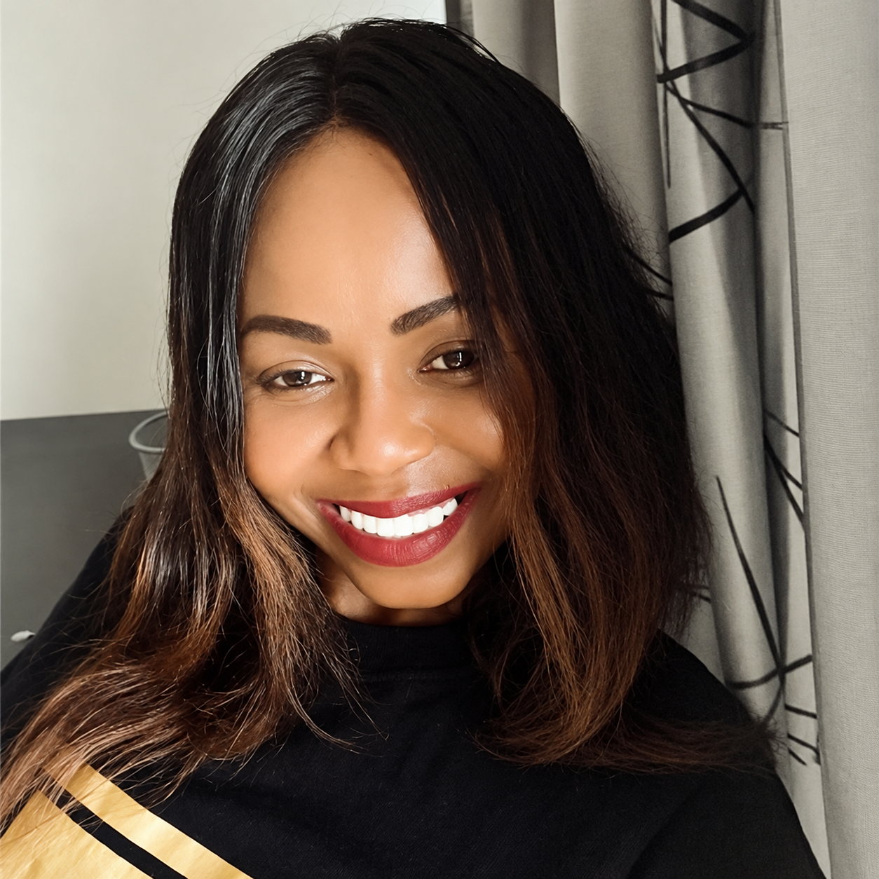 Close-up of a woman with medium-length dark hair, smiling with white teeth, wearing red lipstick, and a black top with gold and black stripes on the shoulder, standing near curtains.