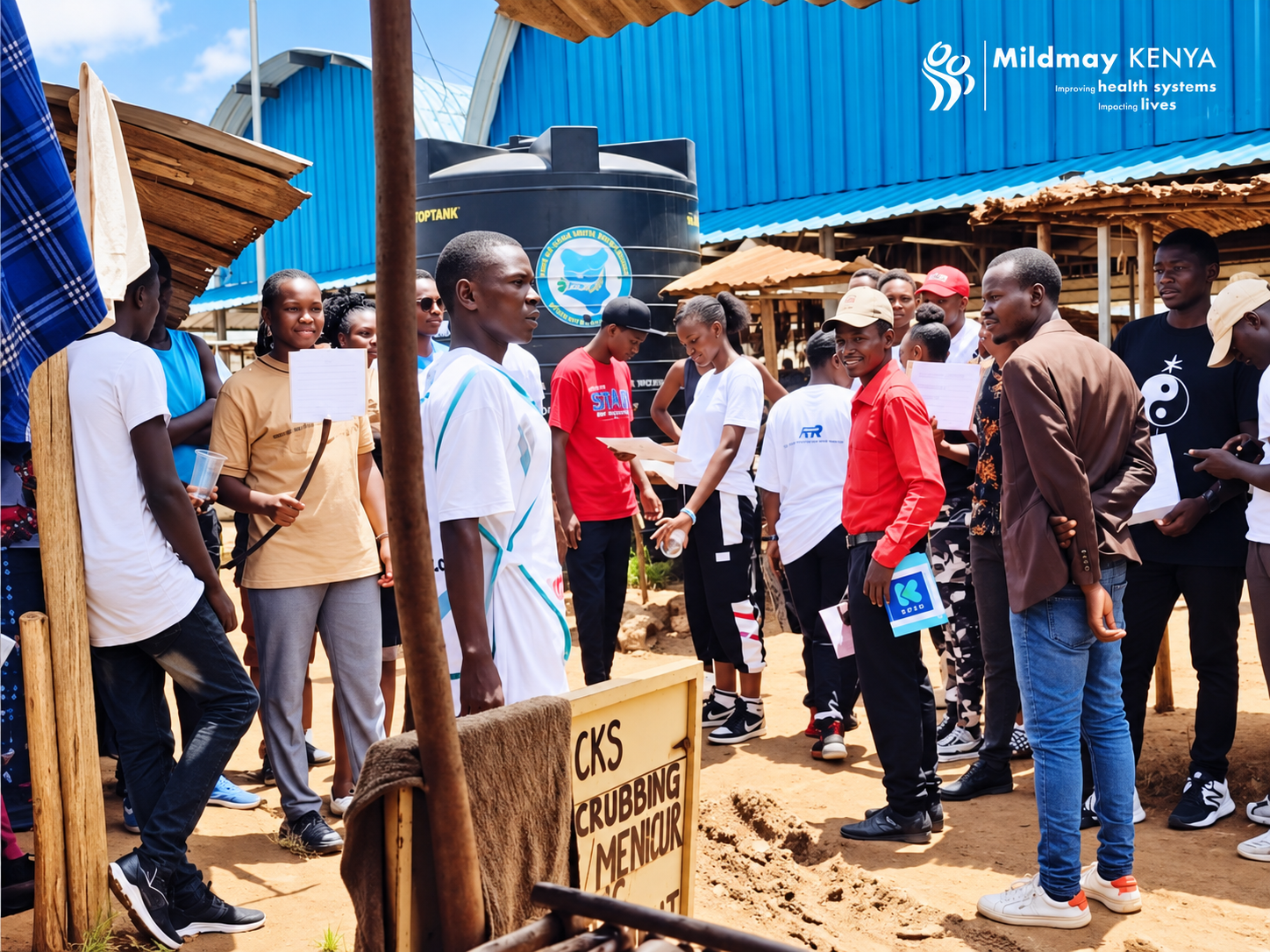 Group of people gathered outdoors at a community event in Kenya, with a blue building and water tank in the background, some holding papers and bags, and a sign indicating a sanitation project.