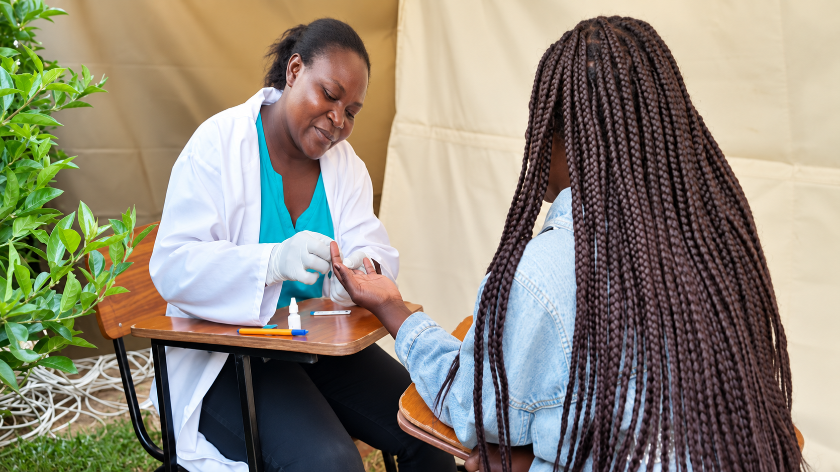 A healthcare professional is performing a finger prick blood test on a woman seated at a table outdoors, with green plants and a beige background.