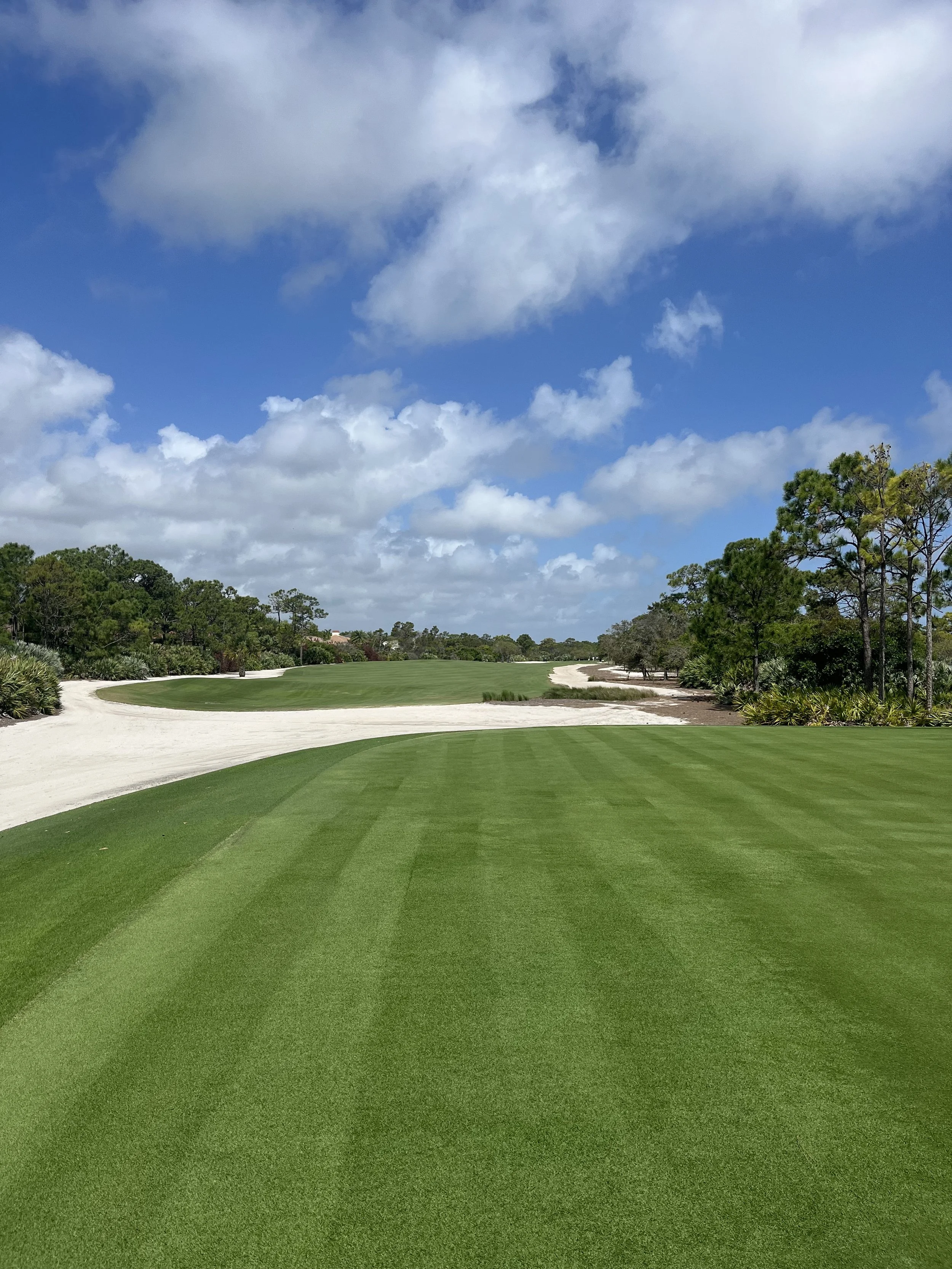 A golf course fairway with green grass, surrounded by sand bunkers and trees under a partly cloudy sky.