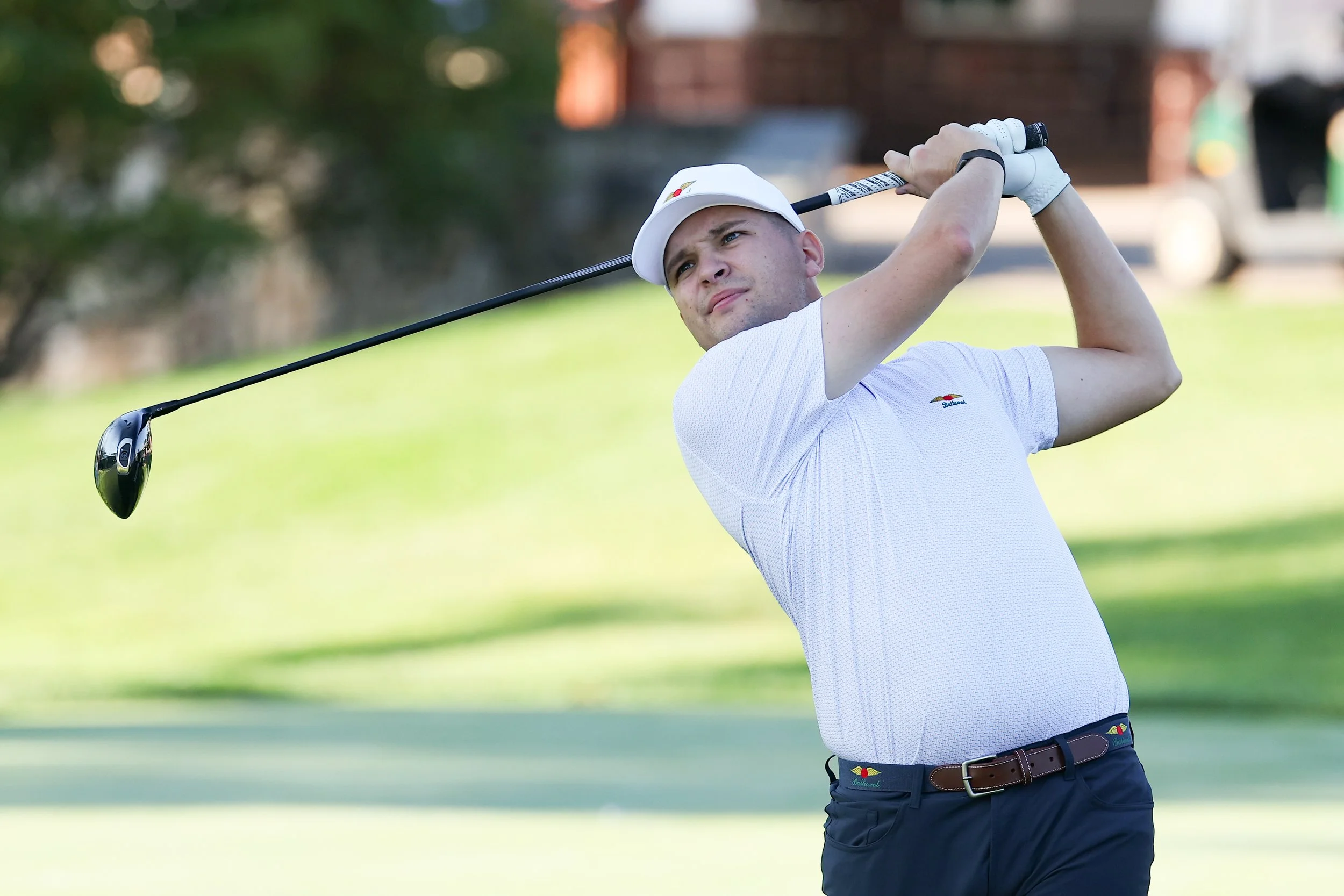 A male golfer is swinging a golf club on a golf course during daytime, wearing a white cap, white shirt, and dark shorts.