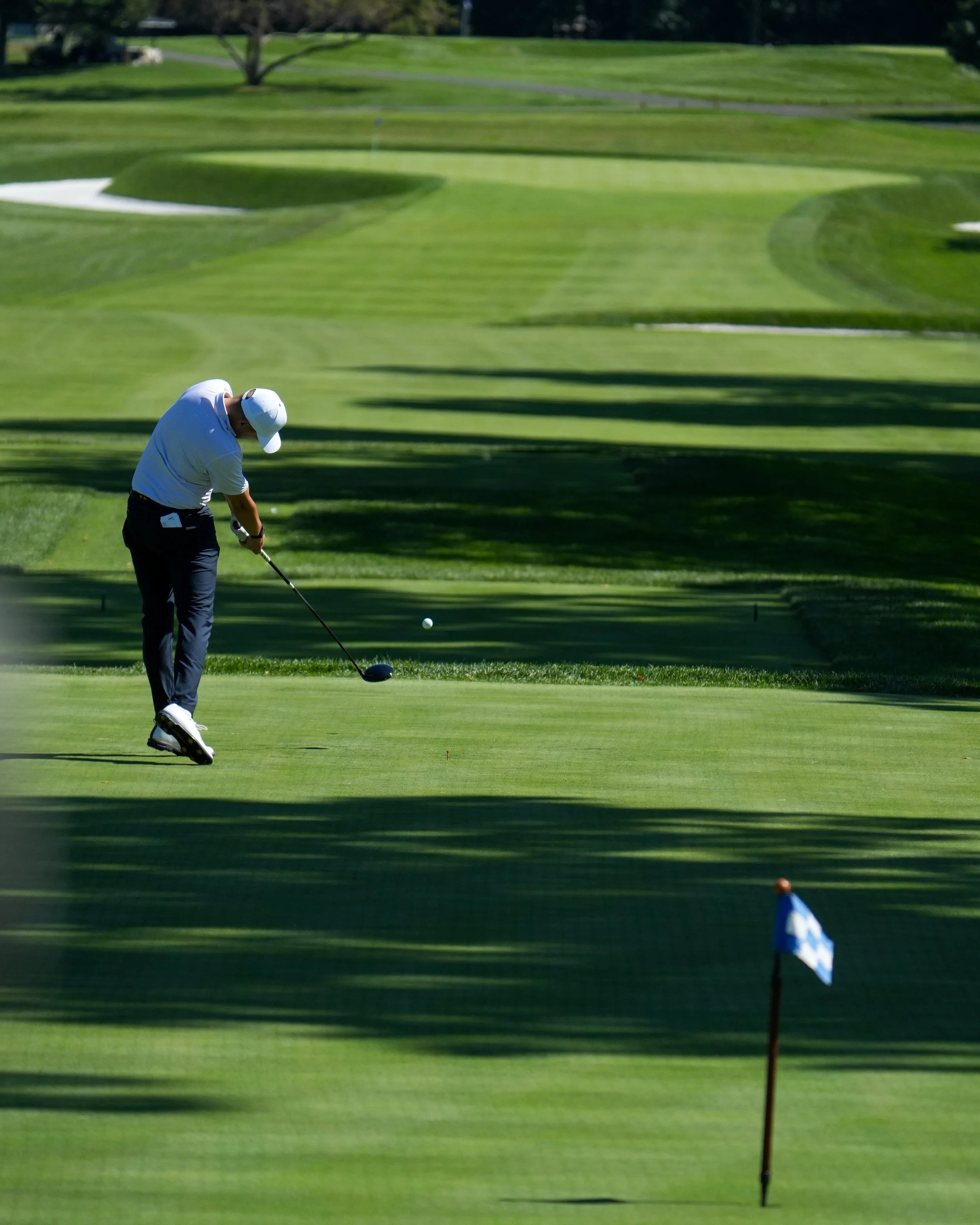 A golfer in a white shirt and black pants swings a club on a golf course. A flag on a small hole marker stands in the foreground, with a well-maintained green and trees in the background.