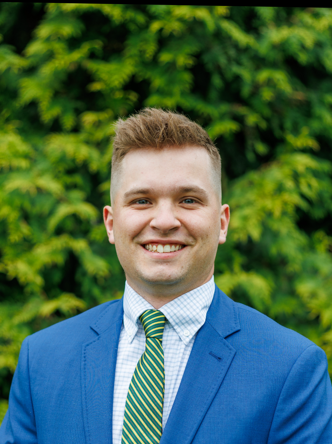 Portrait of a young man smiling outdoors with green foliage in the background, wearing a blue blazer, white checkered shirt, and a striped green and yellow tie.