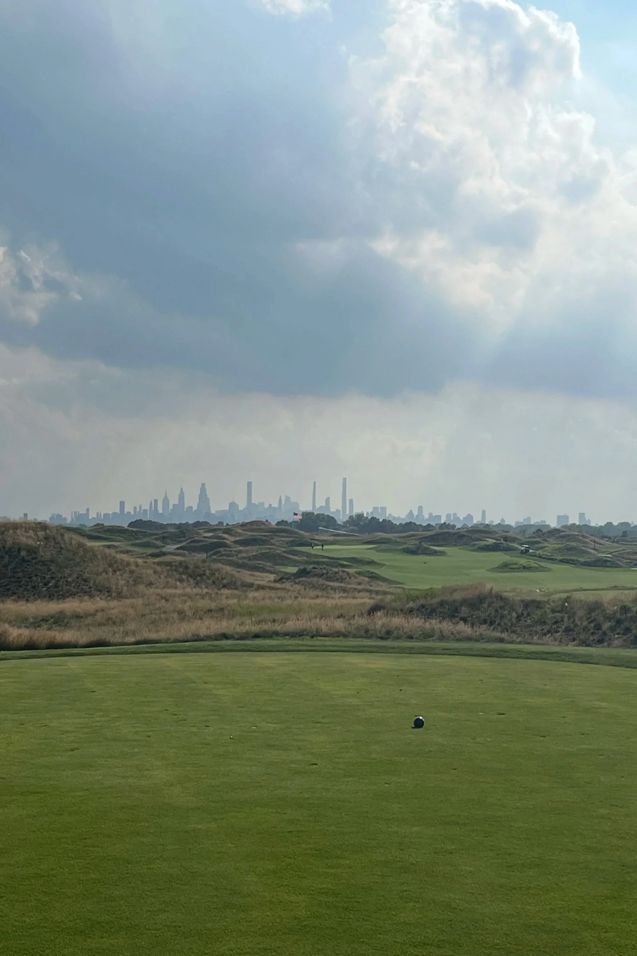 A view of a golf course with a putting green in the foreground, a golf ball on the green, grassy dunes and hills, and a city skyline with tall buildings in the background under a partly cloudy sky.
