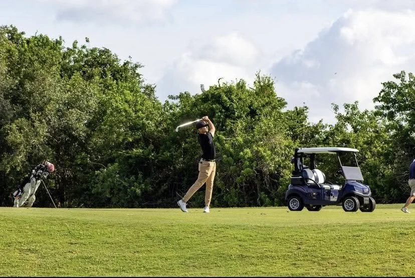 A man in black shirt and khaki pants swinging a golf club on a golf course with lush trees in the background, a golf cart nearby, and a golf bag on a stand at the side.