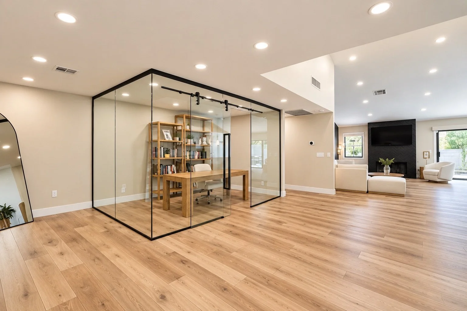 Open-concept living space with hardwood floors, a glass enclosed home office, a beige seating area with a black fireplace, and large windows with sliding doors.