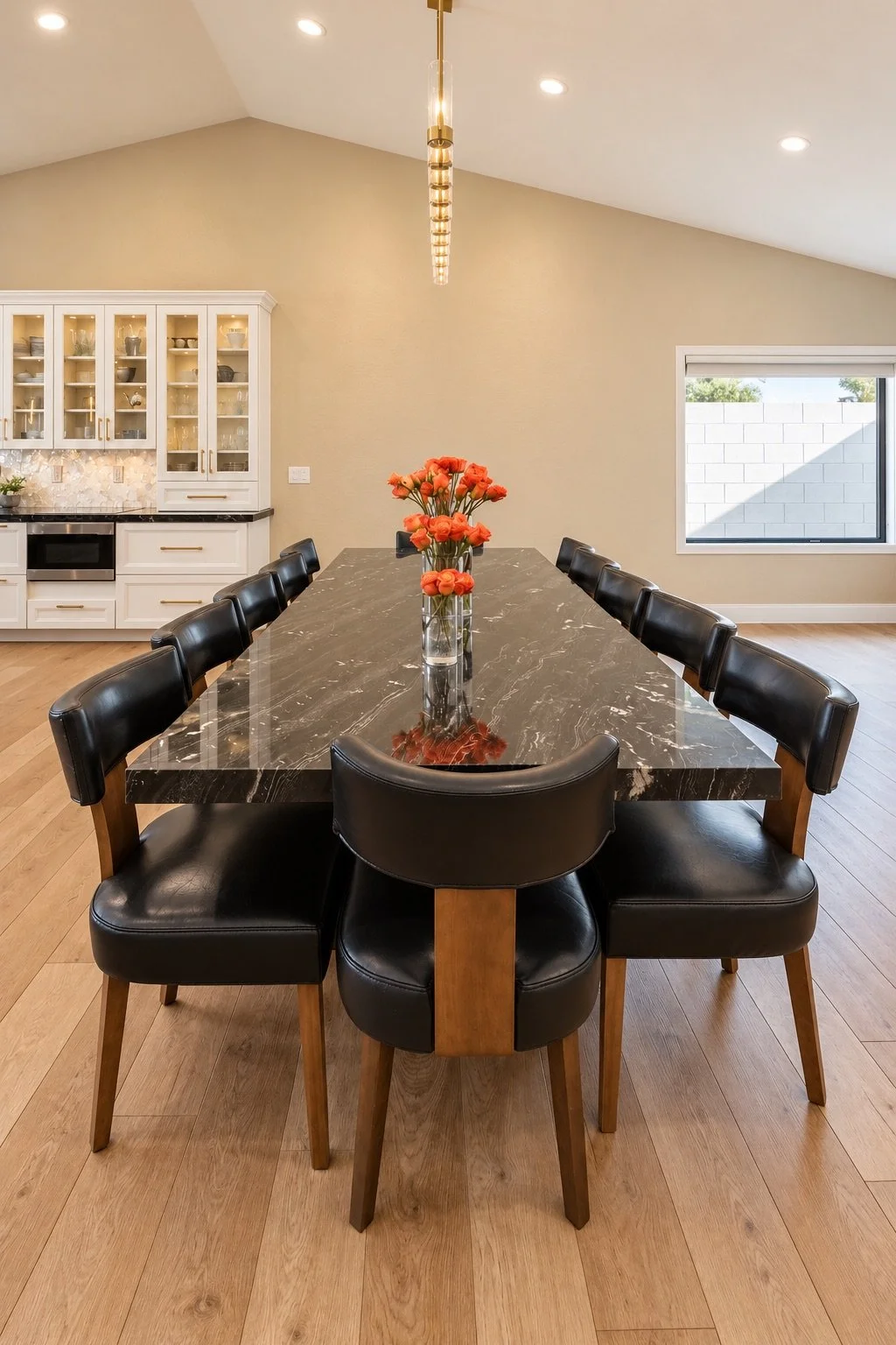 Modern dining room with a black marble table, black leather chairs, a glass vase of orange flowers, a white cabinet with glass doors, and a window with a partially visible brick wall outside.
