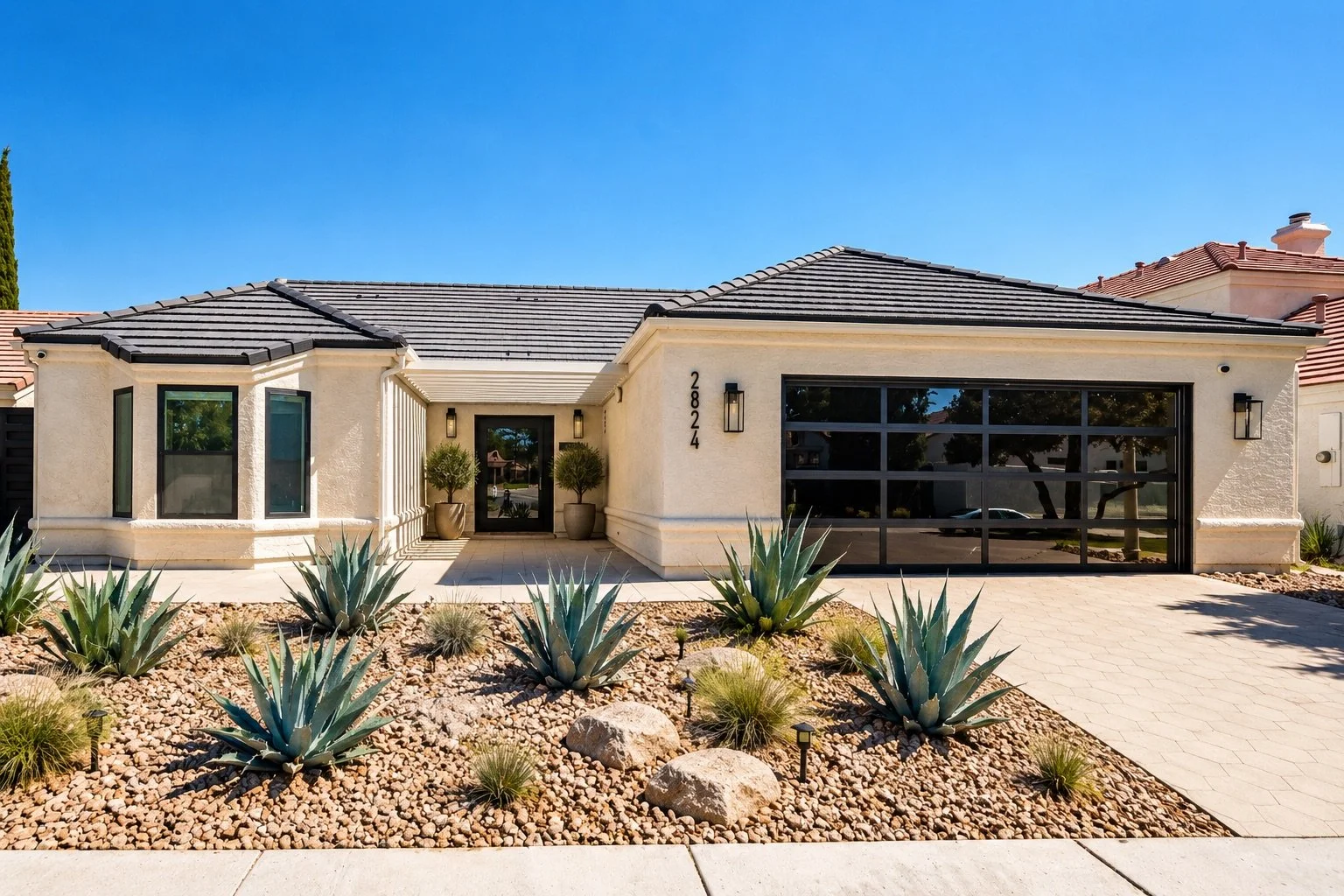Front of a modern house with desert landscaping, large glass garage door, and a front door flanked by two potted plants. The driveway is paved, and the house has a light-colored exterior with a dark tiled roof.