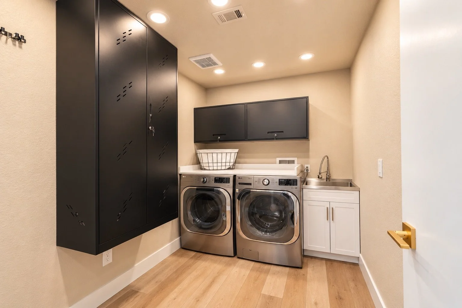 Laundry room with black storage cabinets, a white countertop, a white laundry basket, a washing machine, a dryer, a small white cabinet, a stainless steel sink, and beige walls with recessed ceiling lights.