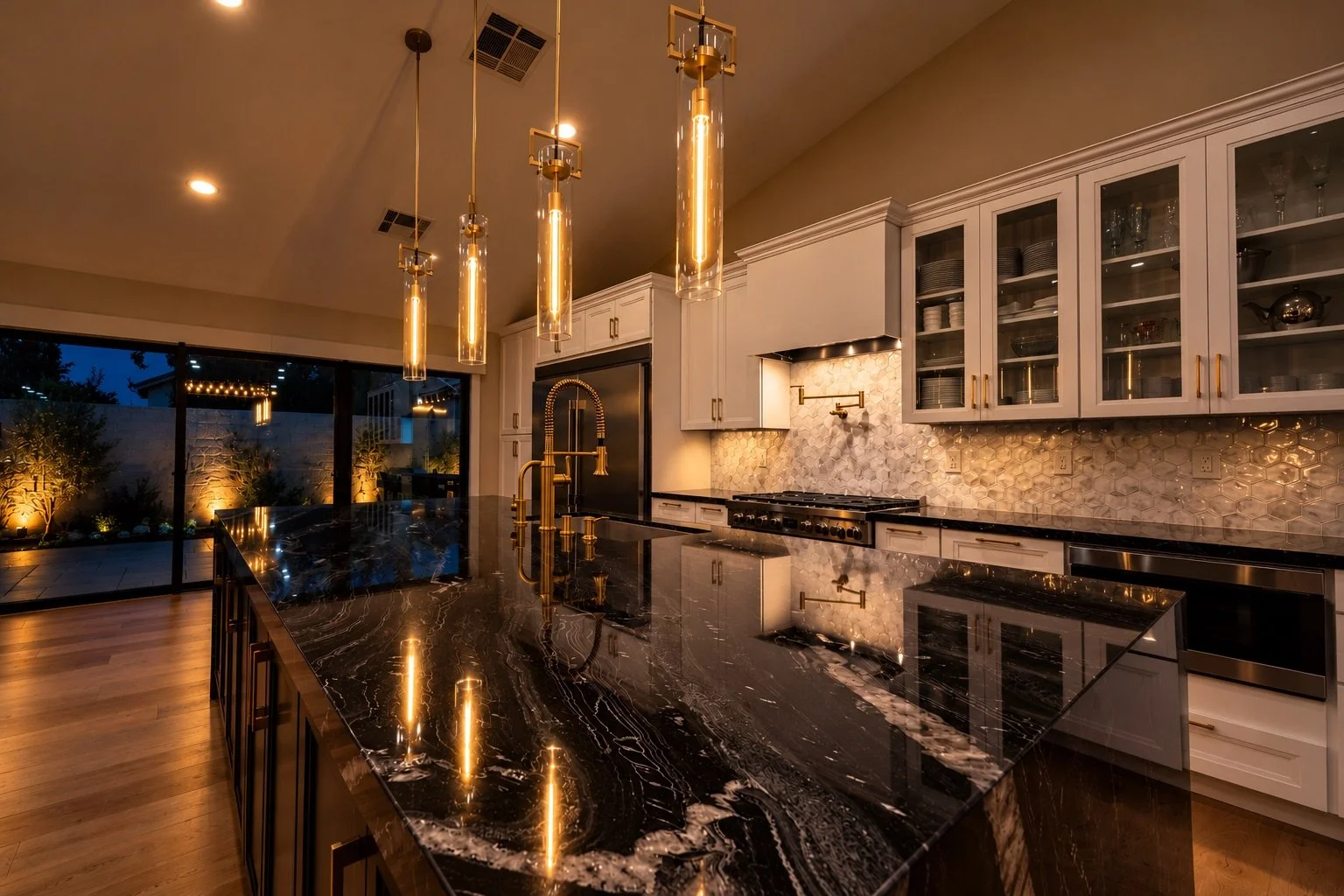 Modern kitchen with black marble island and gold fixtures, pendant lighting, white cabinets, honeycomb backsplash, and sliding glass doors showing outdoor landscaping at night.