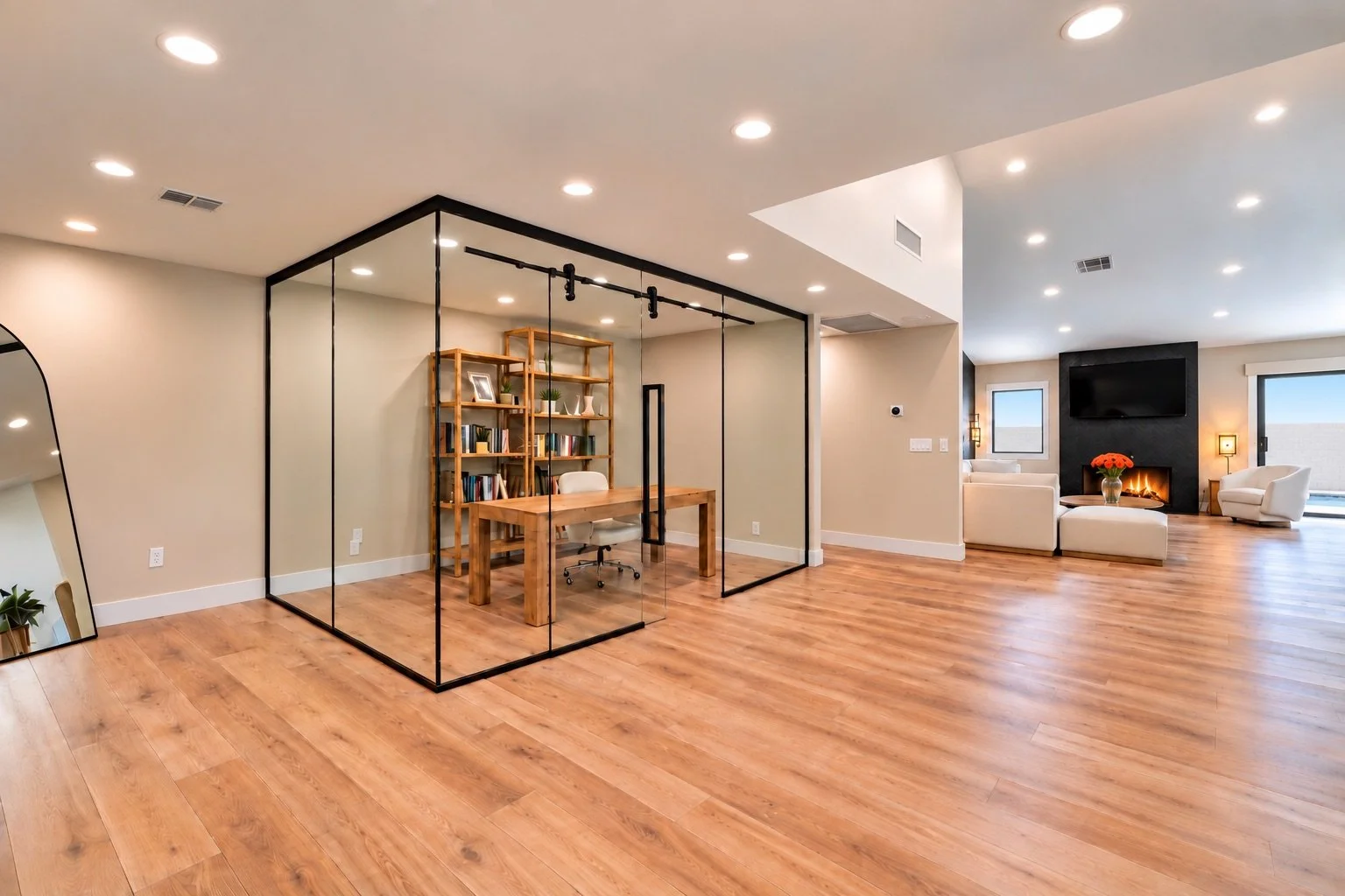 Modern open-concept living room with hardwood floors, a glass-enclosed office with a wooden desk and bookshelf, a black fireplace with a flat-screen TV above, and white seating area with a vase of orange flowers.