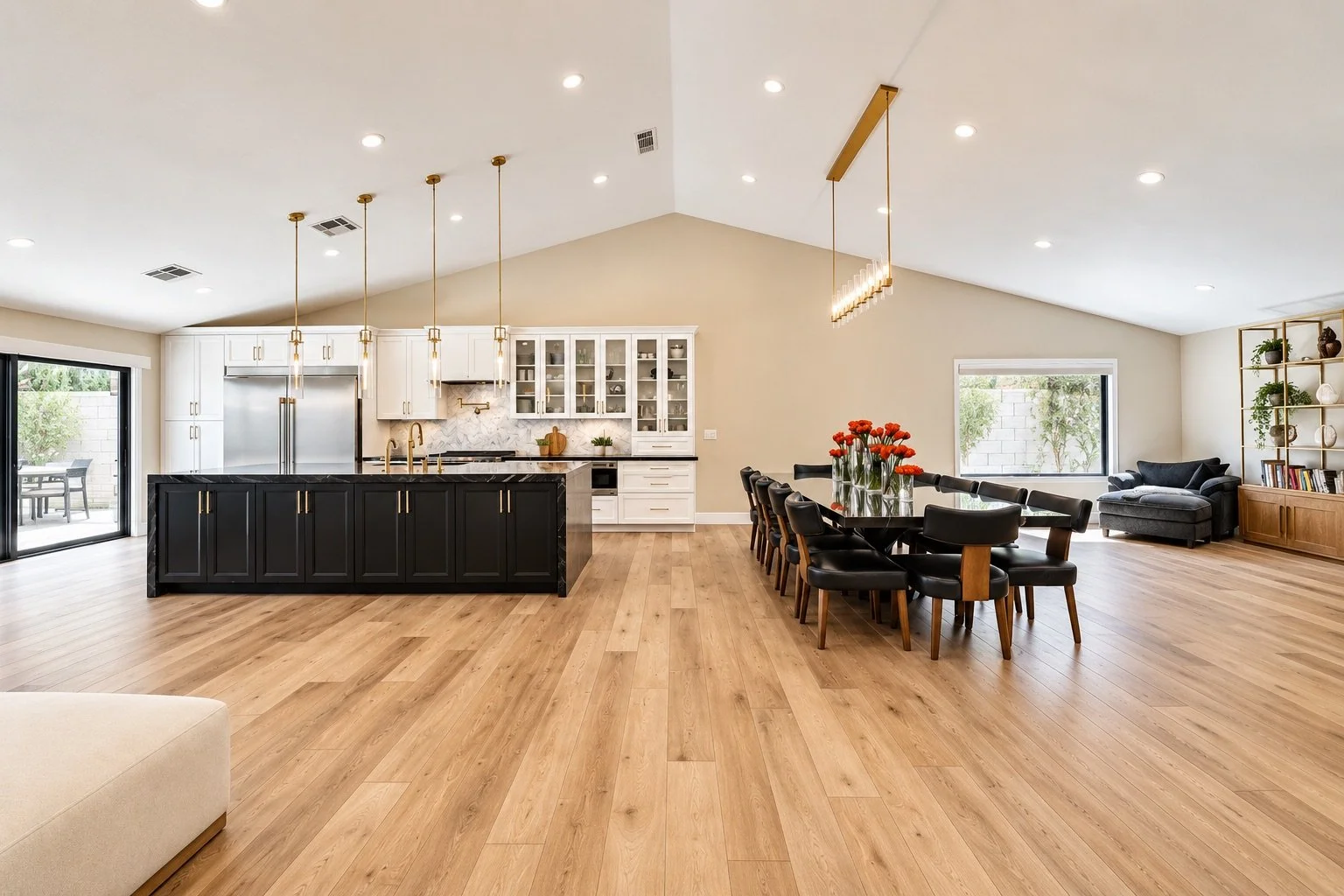 Open-concept living and kitchen area with wooden floors, white ceiling, black dining table with chairs, kitchen with white cabinets, black island, stainless steel appliances, and living space with gray sofa and shelves.