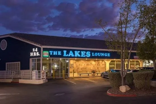 Night view of The Lakes Lounge with a bright blue sign, some cars parked outside, and trees in the foreground.
