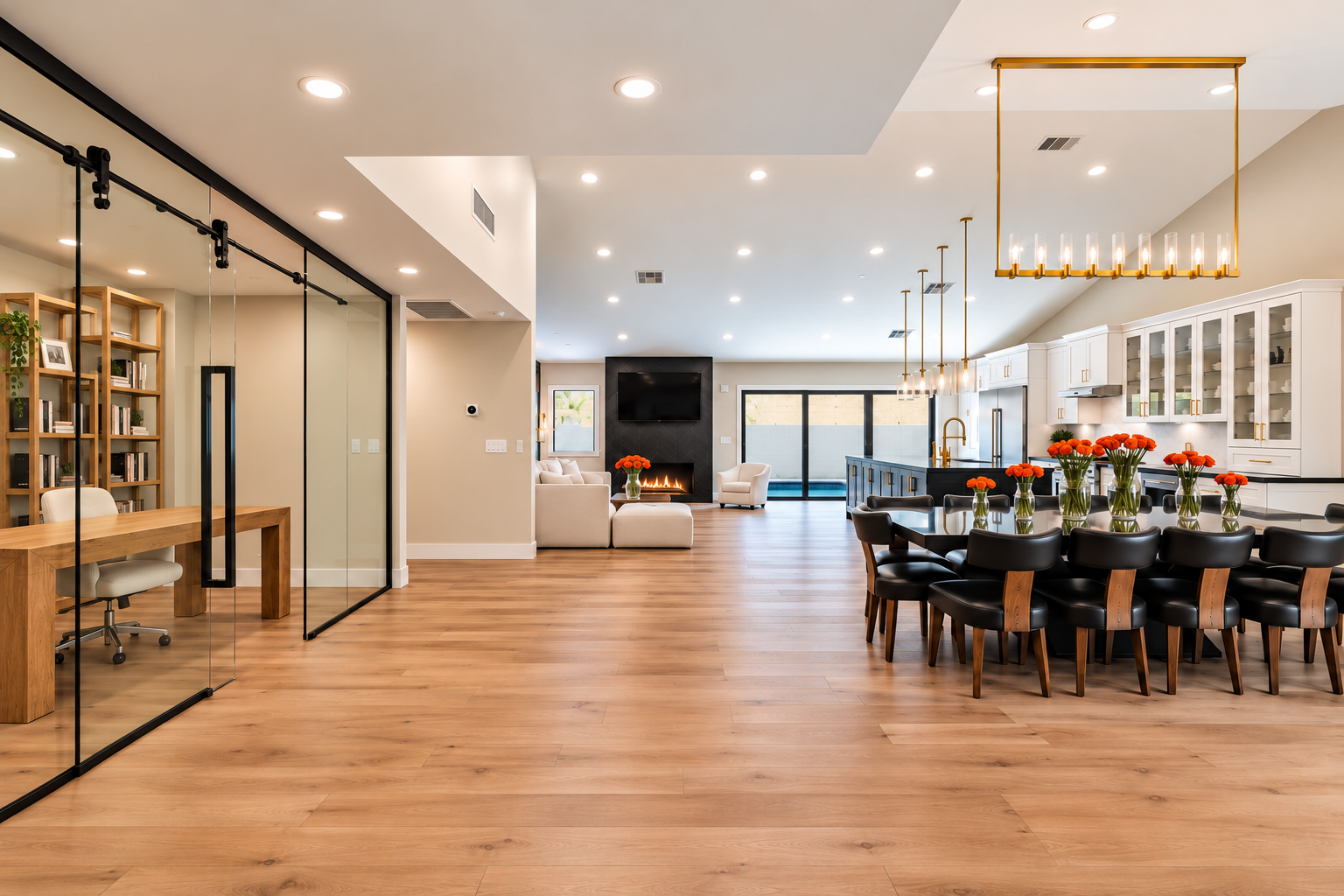 Open-concept living room and kitchen with hardwood floors, white cabinets with glass fronts, black countertops, and a large dining table with black chairs and orange flowers. There is a fireplace, sliding glass doors, and modern lighting fixtures.