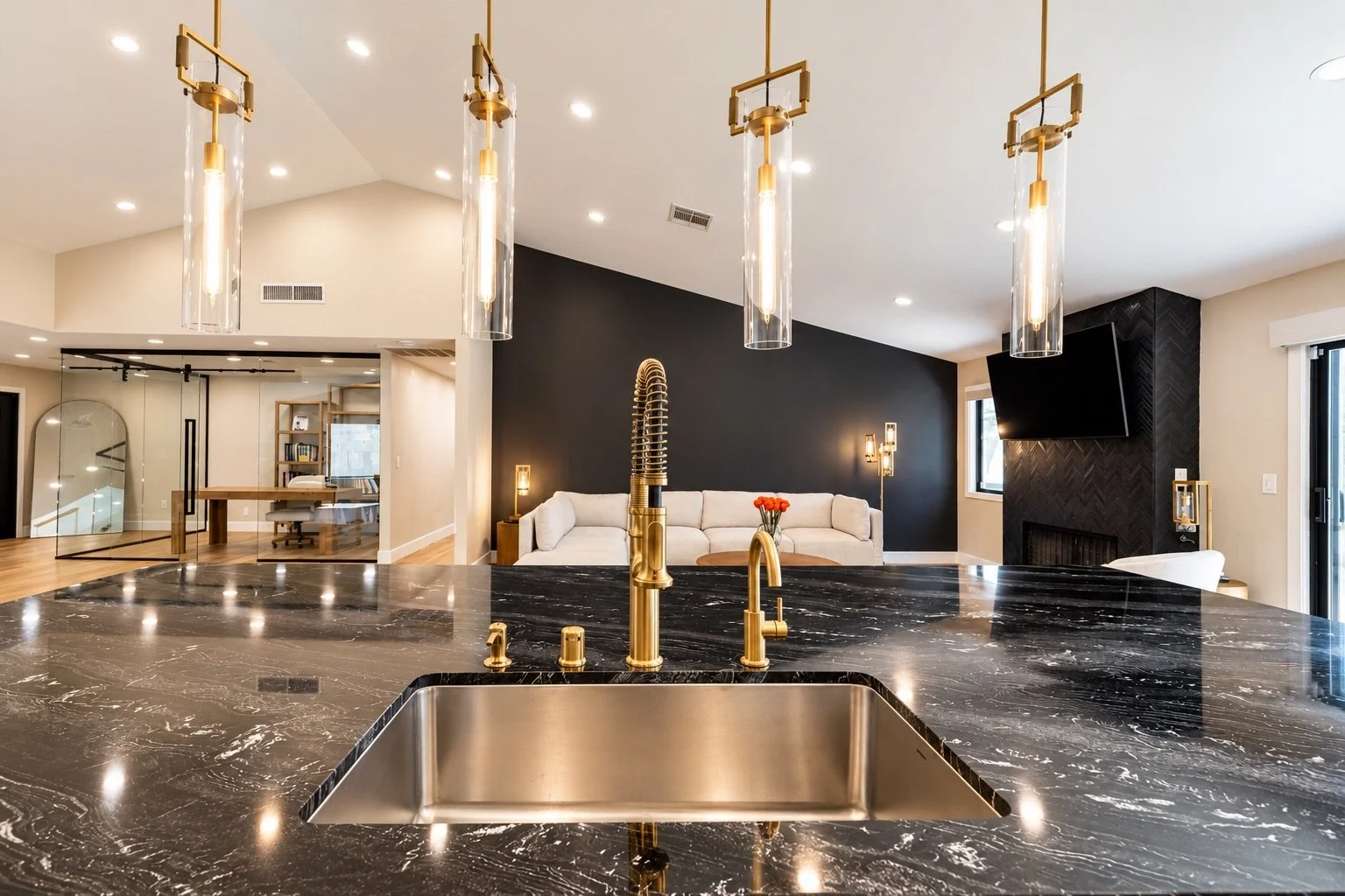 Modern kitchen with black marble island, gold faucet, and sleek lighting, overlooking a living area with white sofa and black accent wall.