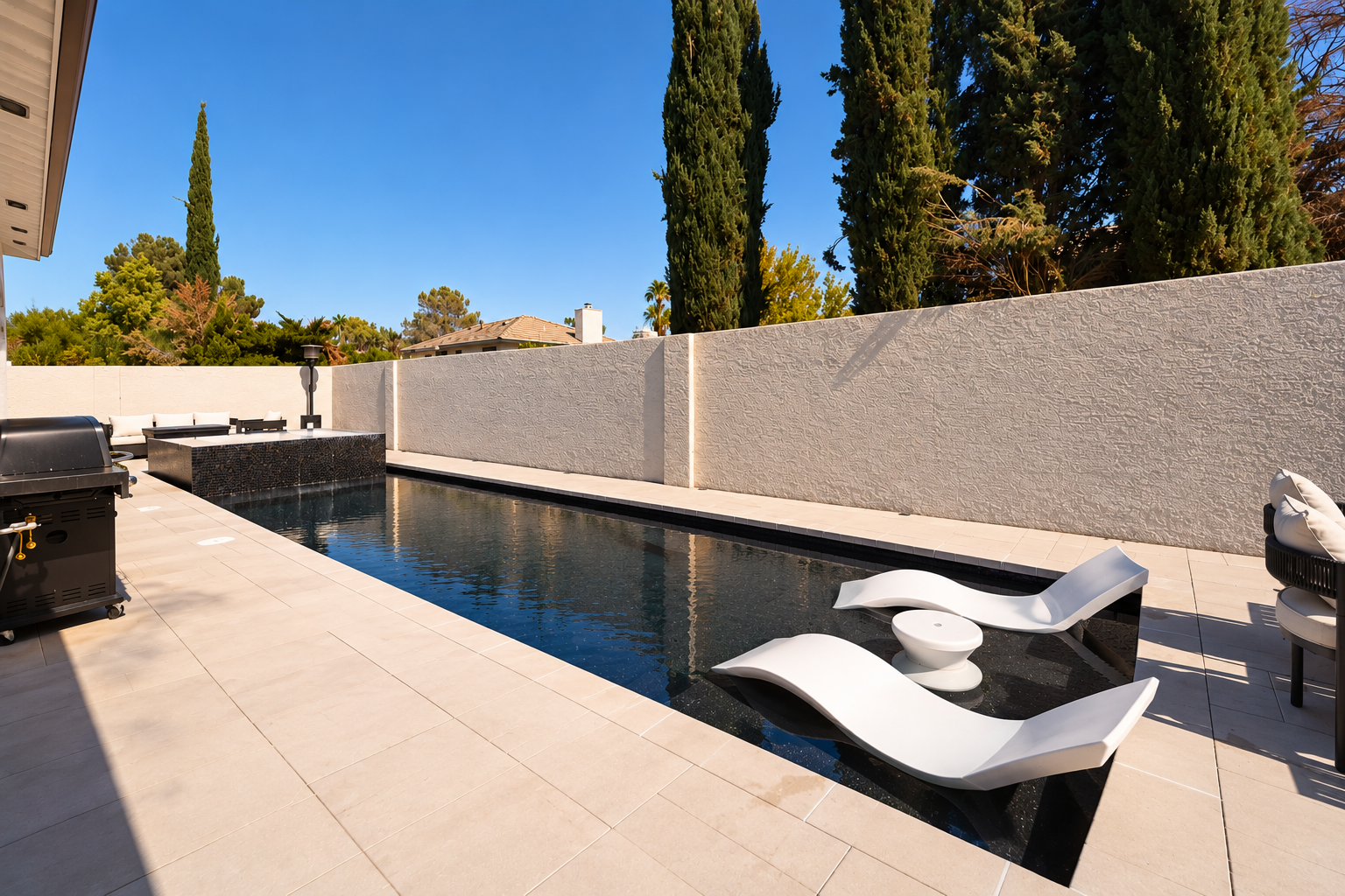 Modern backyard with a narrow black swimming pool, white lounge chairs partially submerged in water, white walls, and tall green trees against a blue sky.