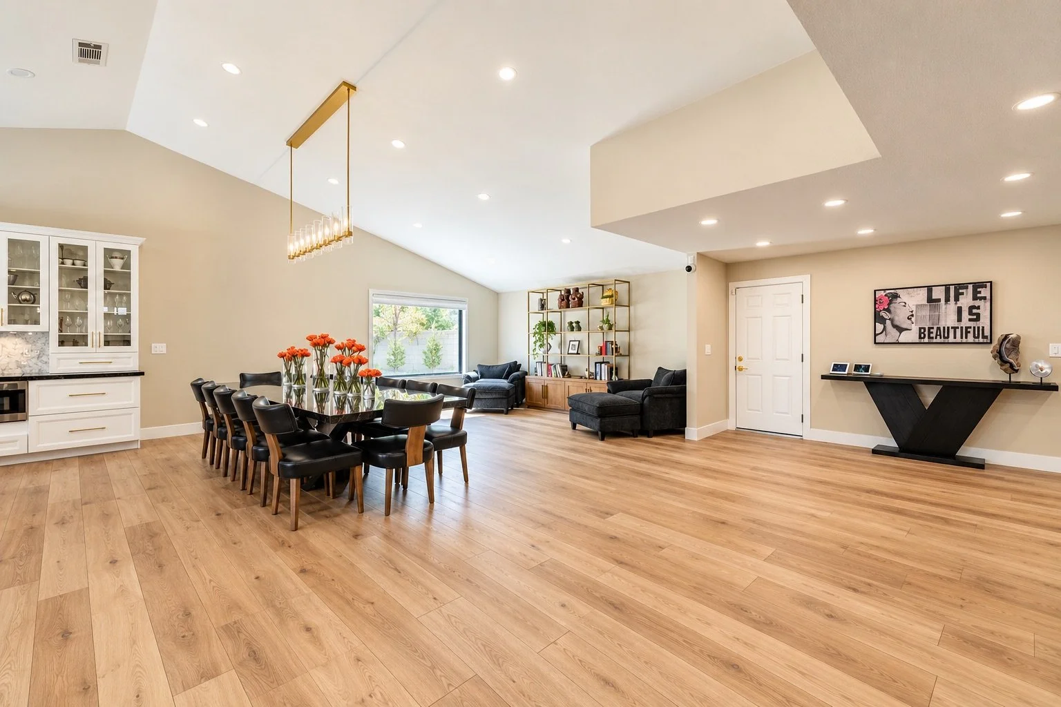 Open-concept living and dining room with hardwood floors, white walls, a large dining table with 12 black chairs, a kitchen area with white cabinets, a window, and a seating area with black sofas and a bookshelf.