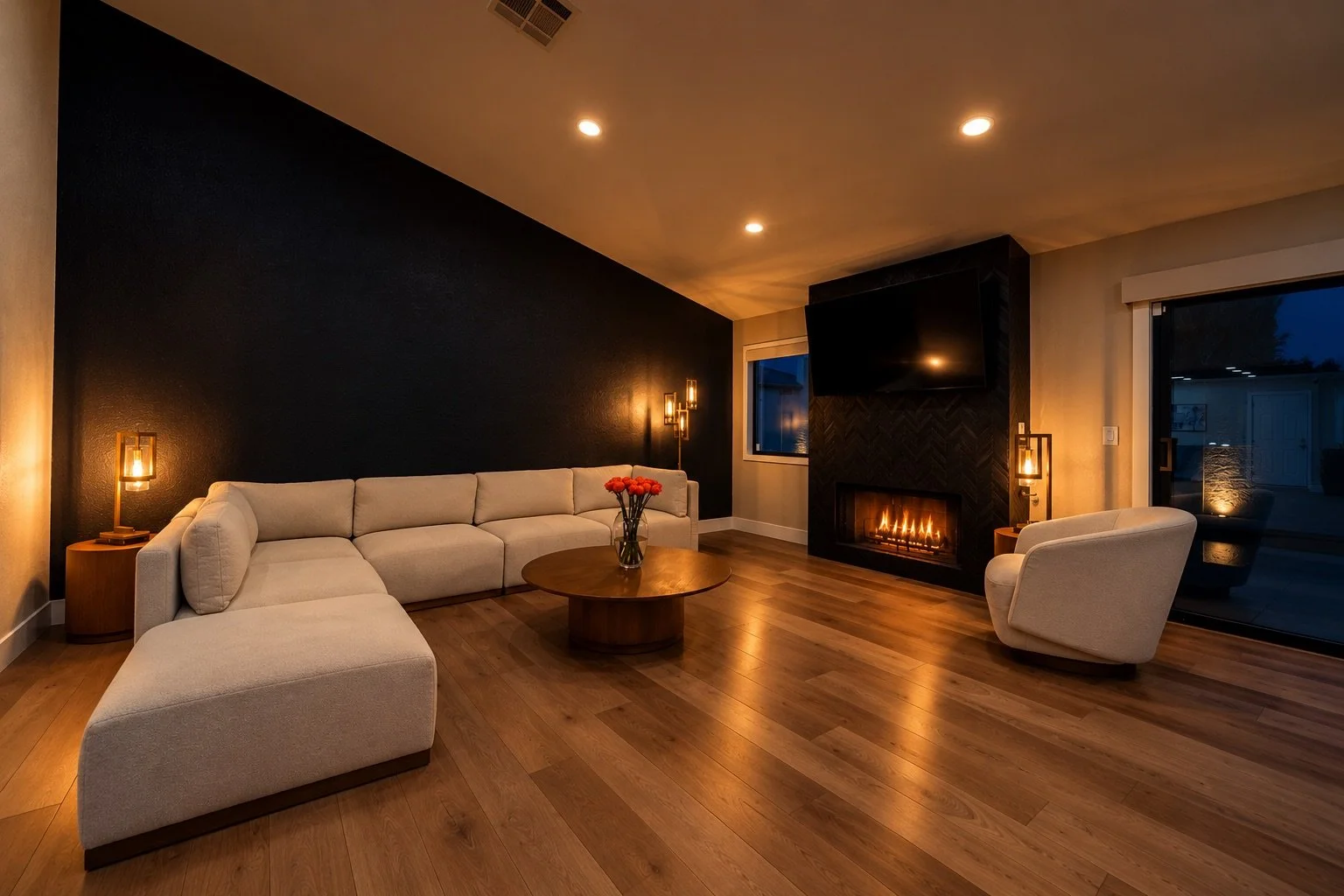 Living room with a black accent wall, beige sectional sofa, white armchair, wooden coffee table with flowers, fireplace, wall-mounted TV, and sliding glass doors, with warm lighting and hardwood floors
