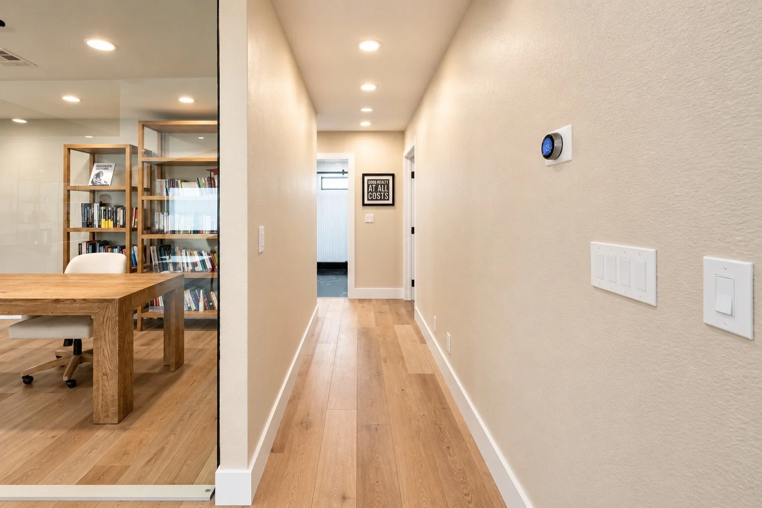 Residential hallway with beige walls and wooden flooring, featuring a thermostat on the right wall, and a sign at the end of the hallway that reads 'Good Realty at All Costs'.