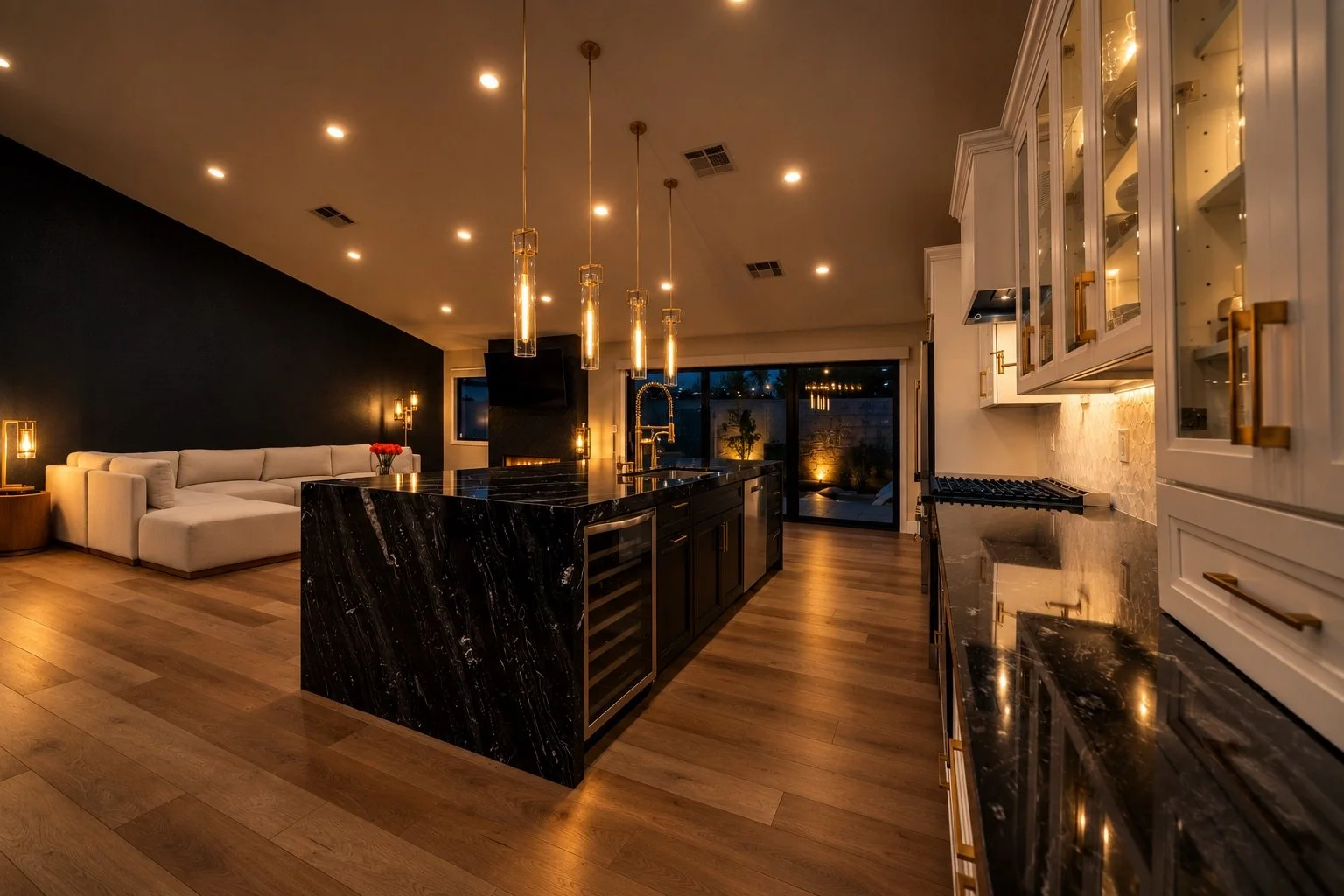 Modern kitchen with black marble island, white cabinetry with gold hardware, wooden floors, and warm ambient lighting, open to living area with a sofa and large window.