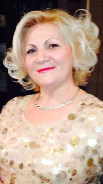 A woman with blonde, curly hair and red lipstick wearing a light-colored top with gold sequins, and a pearl necklace, smiling at the camera.