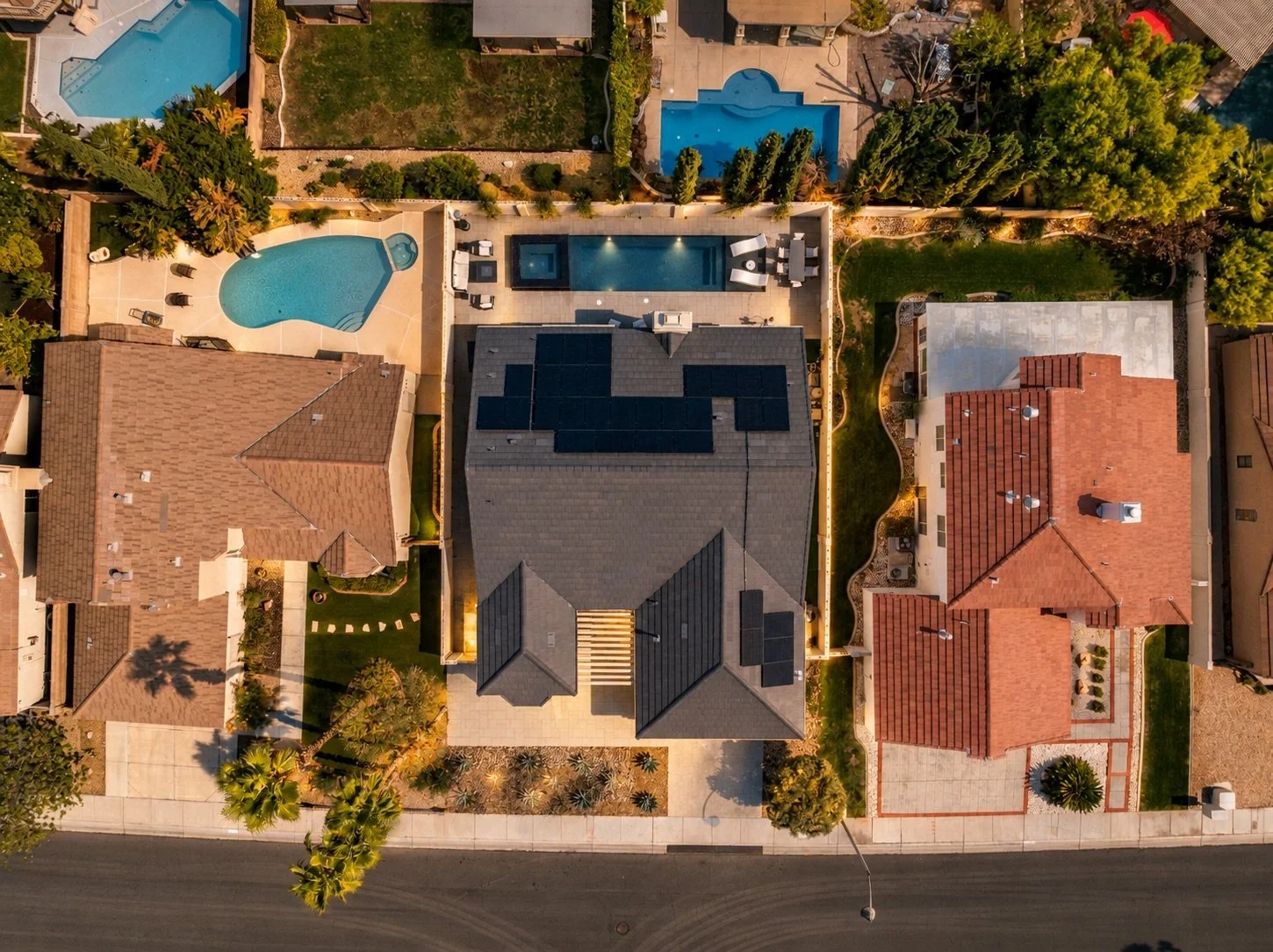 An aerial view of a residential backyard with a pool, patio, and backyard amenities, surrounded by neighboring houses and trees.