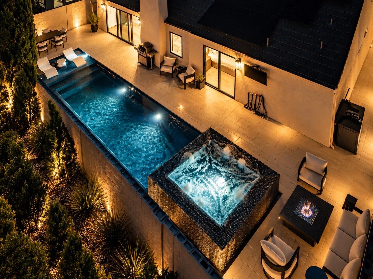 A backyard at night with a lit swimming pool, lounge chairs, patio seating, and a hot tub. Surrounding trees and outdoor lighting create a cozy atmosphere.