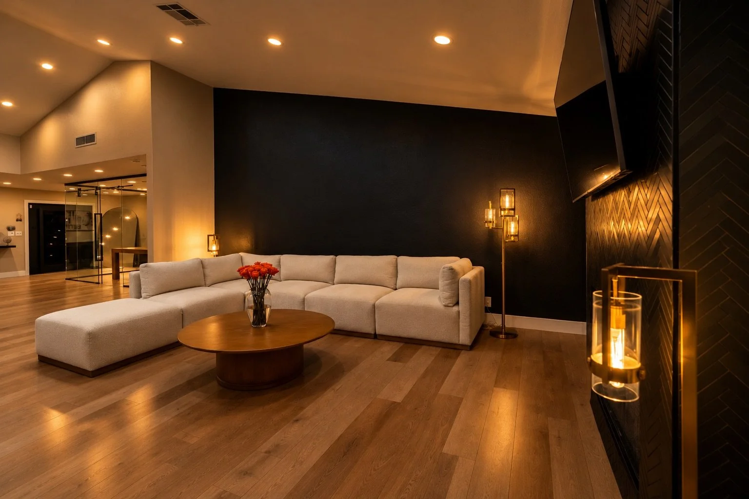 Modern living room with a large beige sectional sofa, a round wooden coffee table with a vase of red flowers, a black accent wall, and warm lighting, including candle-like fixtures.