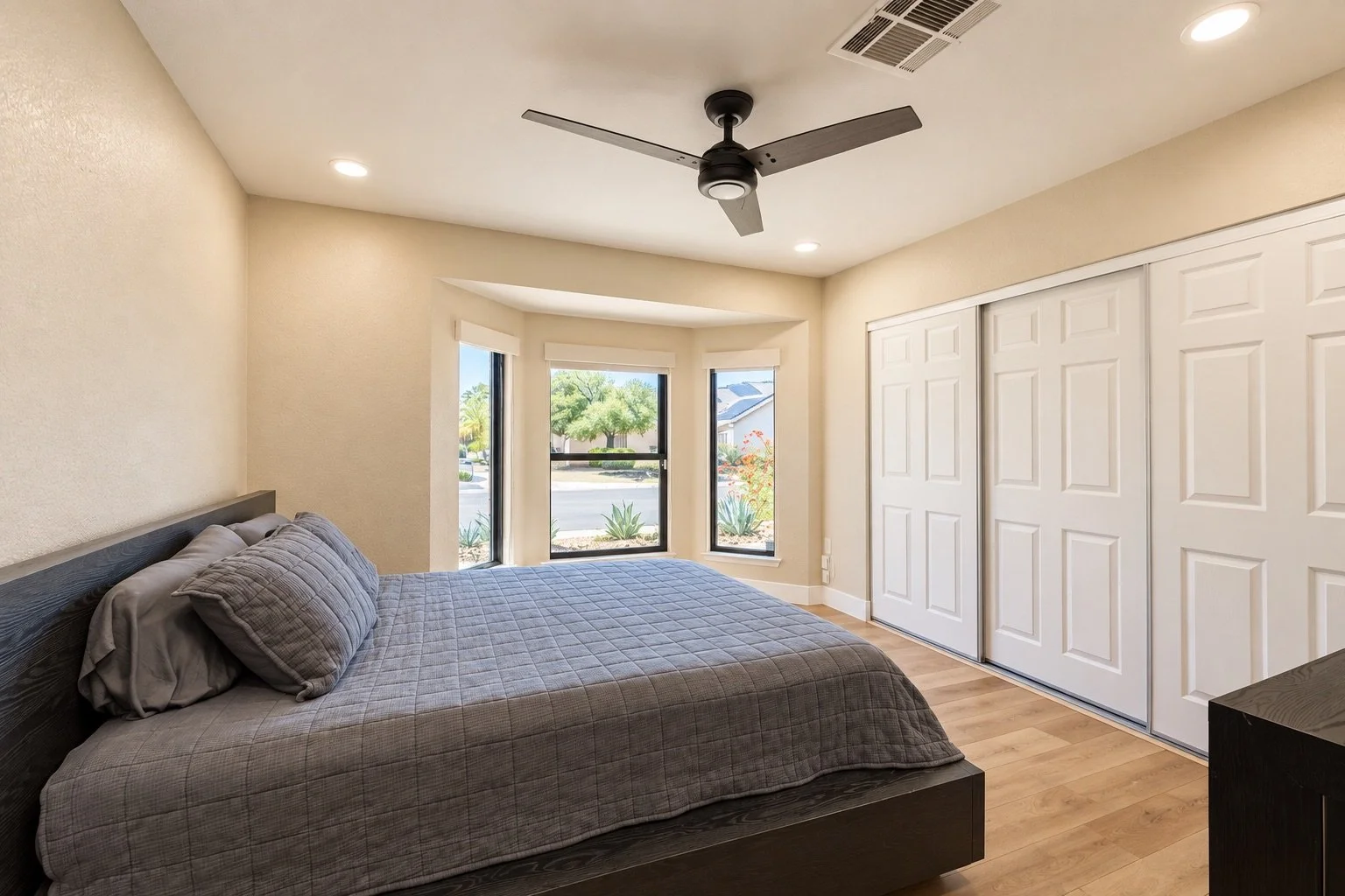 A bedroom with a bed covered in a gray checkered quilt, beige walls, and a ceiling fan. It has three large windows with white blinds, showing trees and a street outside. There are white closet doors on one wall and a wooden nightstand.
