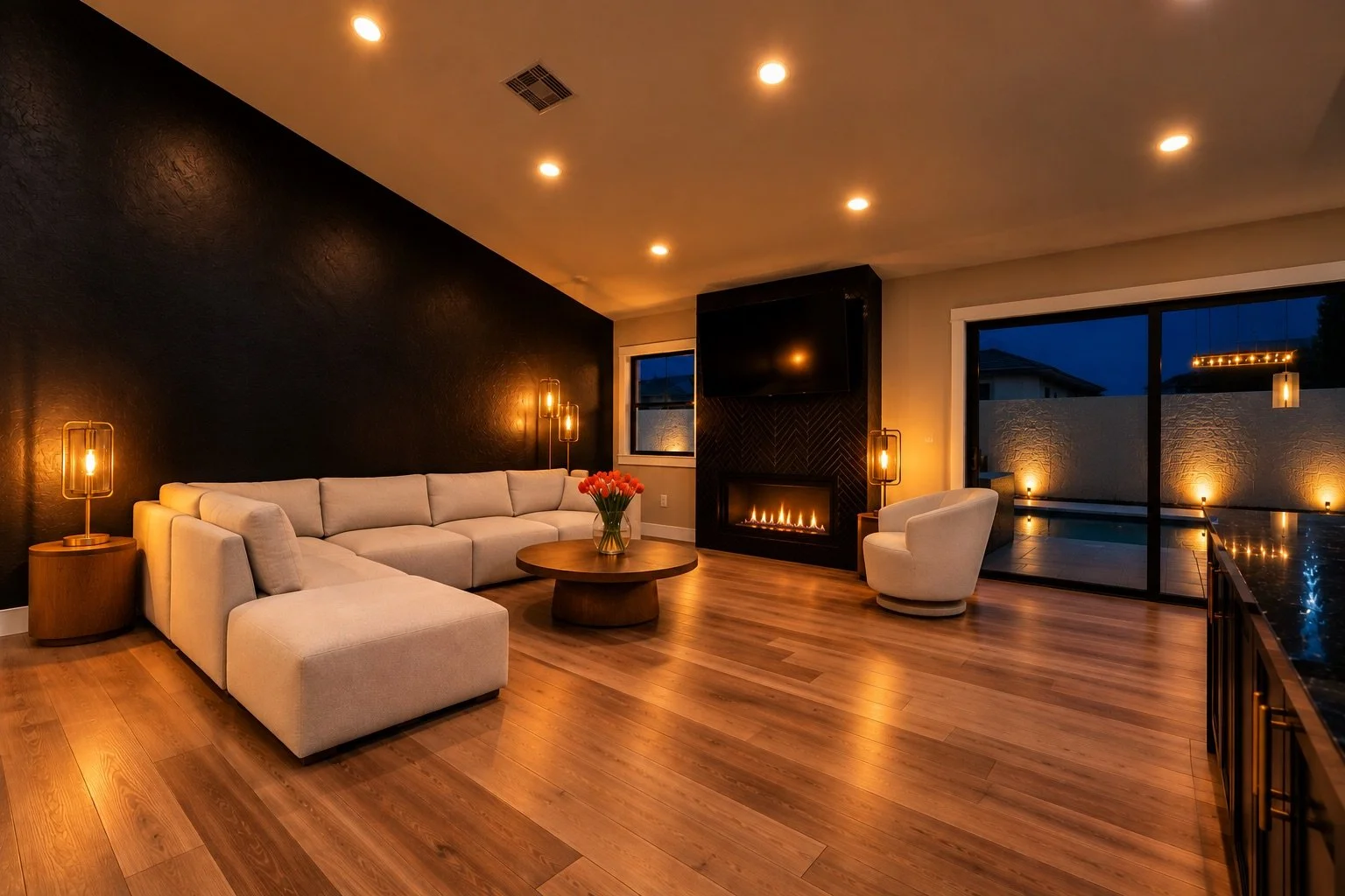 Living room with a large beige sectional sofa, a white armchair, a circular coffee table with a flower vase, a fireplace, a flat-screen TV, sliding glass door, hardwood floors, and modern lighting, evening