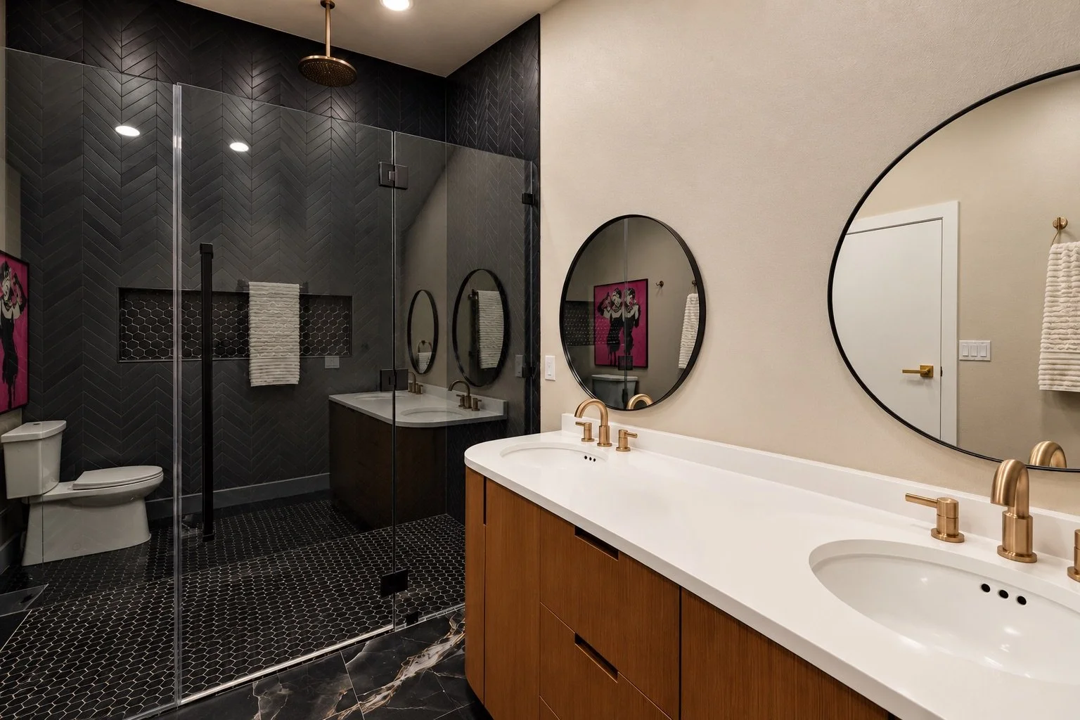 Modern bathroom with dual sinks featuring gold faucets, large oval mirrors, and a black-tiled walk-in shower with a glass door and a white towel hanging on a black towel bar. Artwork on the wall.