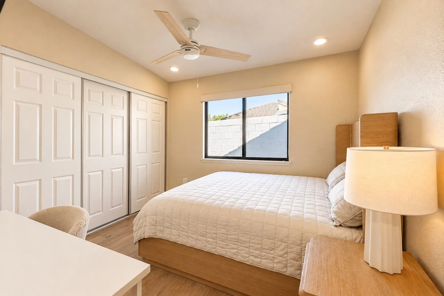 A bedroom with beige walls, a window with a view of a gray brick wall, a wooden bed frame with a white quilt, a wooden nightstand with a cream-colored lamp, a ceiling fan, and closet with white sliding doors.